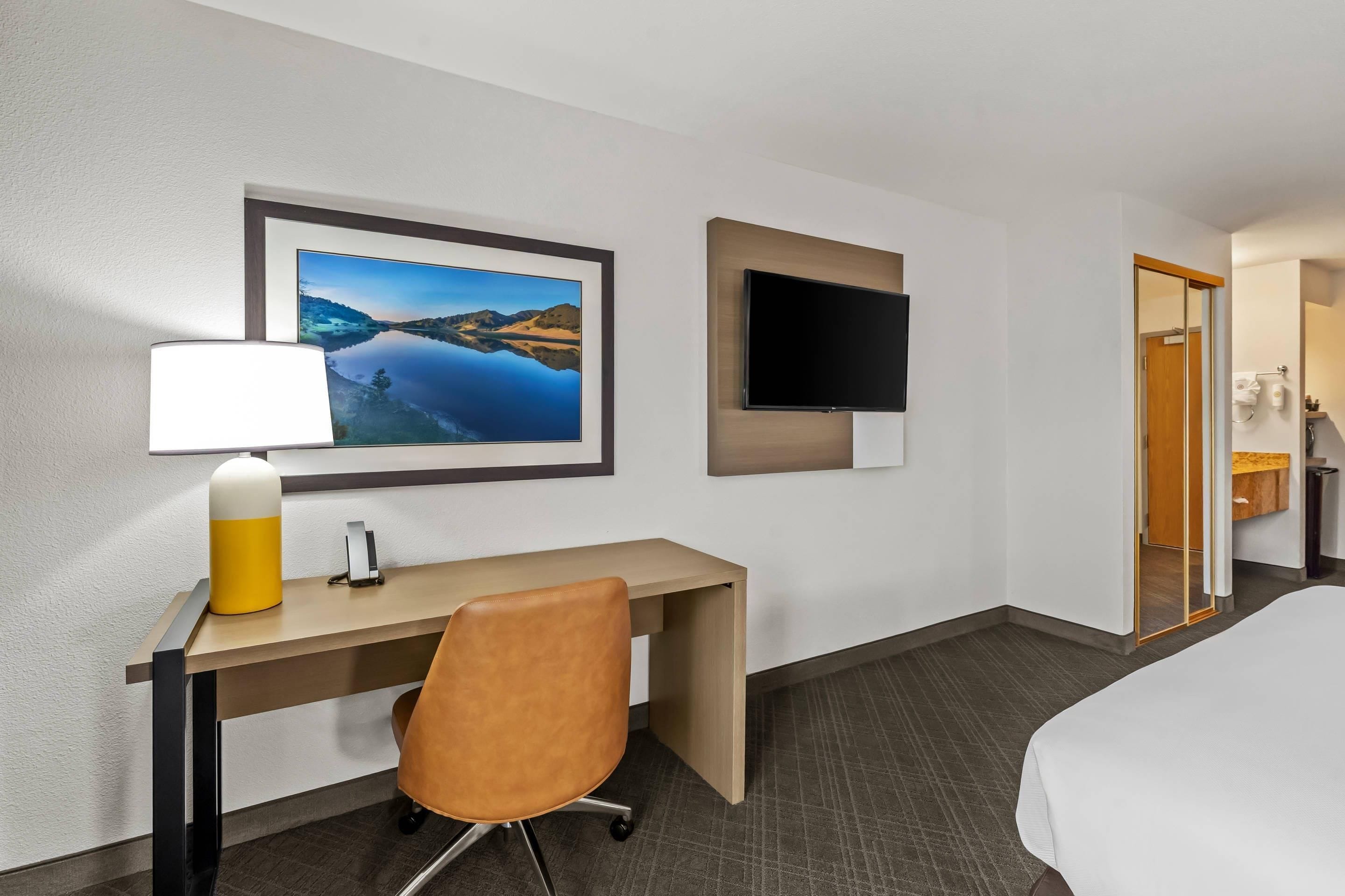 A hotel room with a bed, a desk with a chair, a flat-screen TV, a framed picture on the wall, and a bathroom door.
