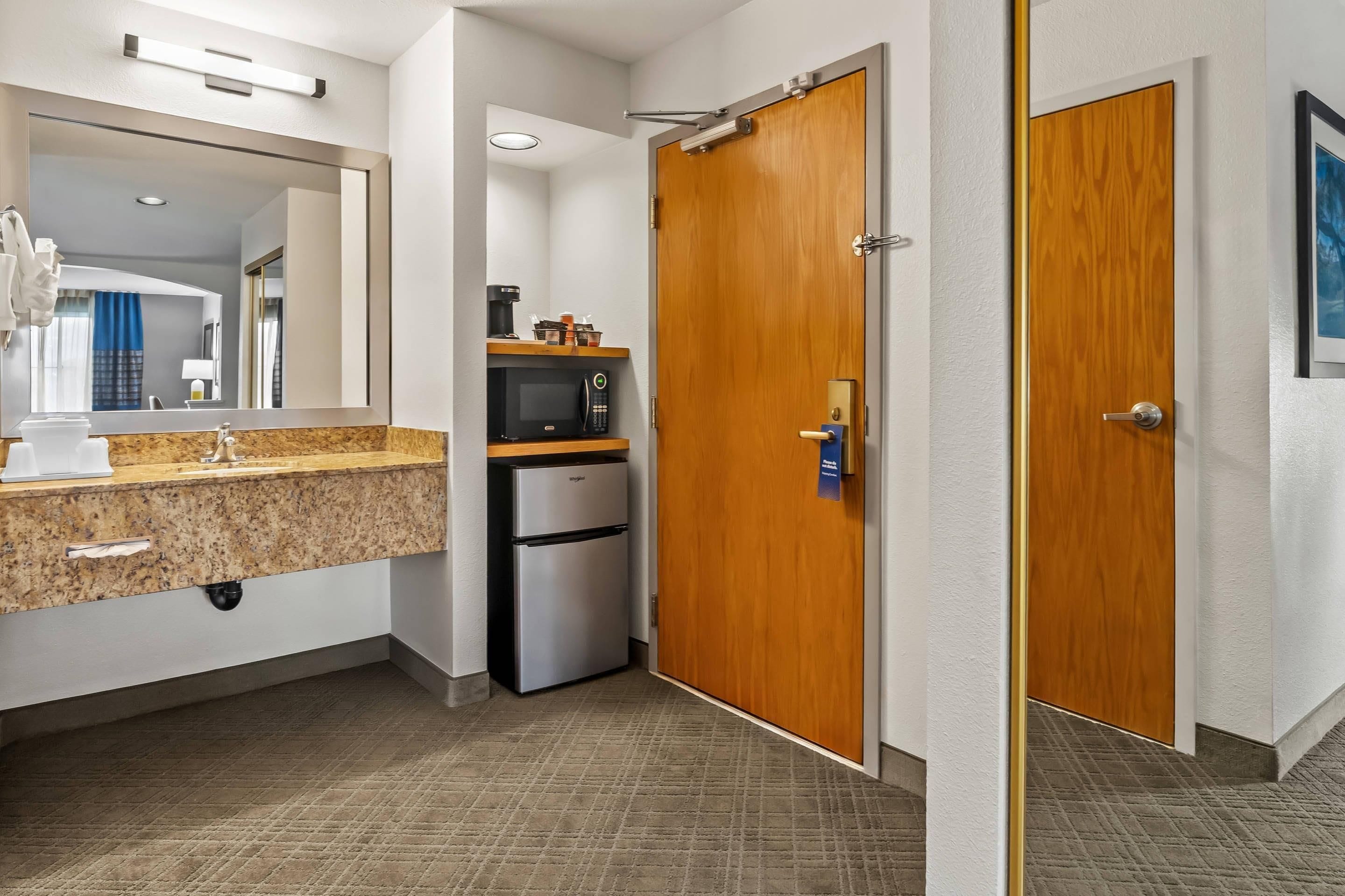 A hotel bathroom with a sink, mirror, and a refrigerator.