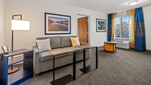 A hotel room with a sofa, a desk, a lamp, framed pictures on the wall, a window with curtains, and an air conditioning unit. - Comfort Inn South San Jose / Morgan Hill (Morgan Hill)