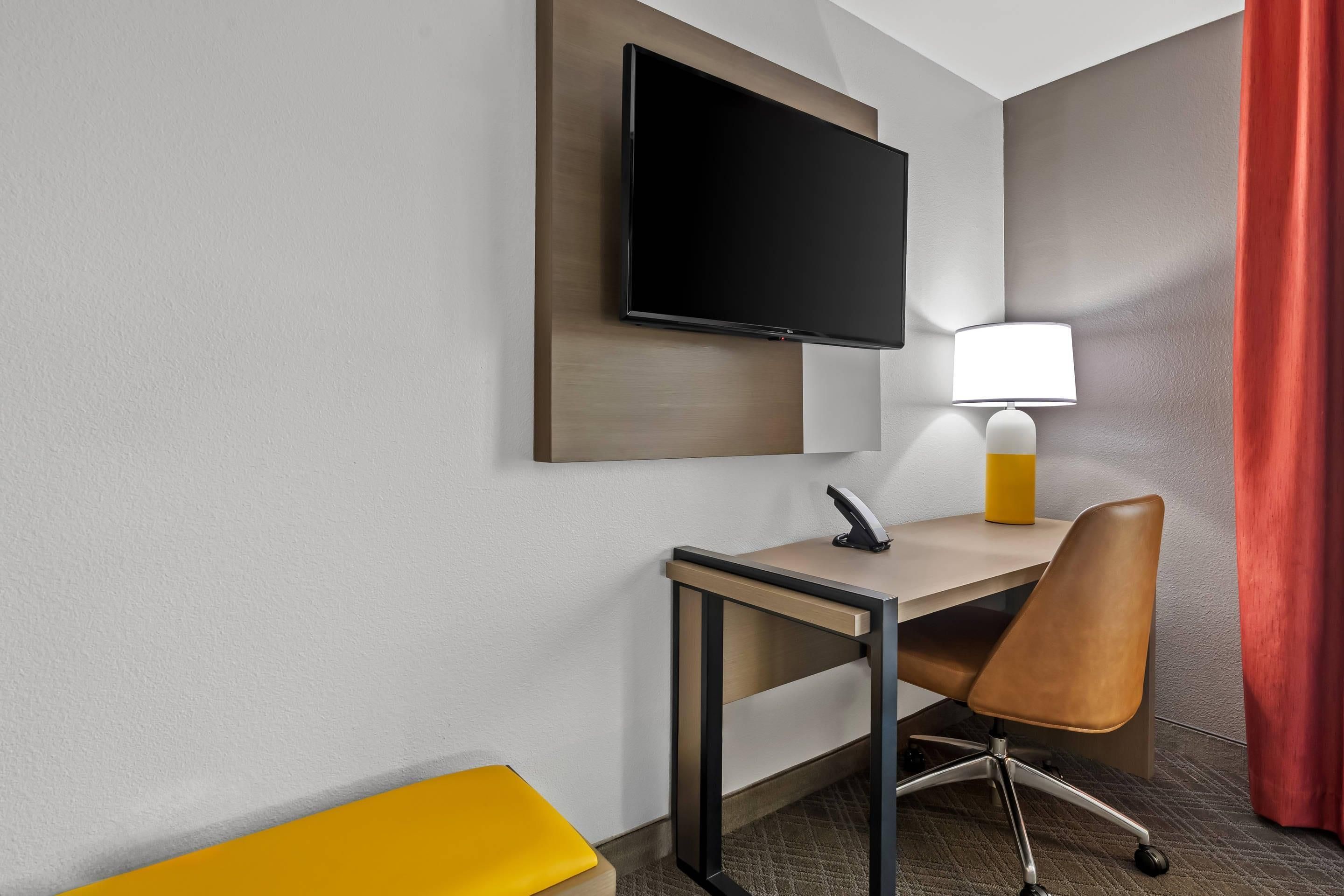 A hotel room with a flat-screen TV mounted on the wall, a desk with a chair, a lamp, and a red curtain.