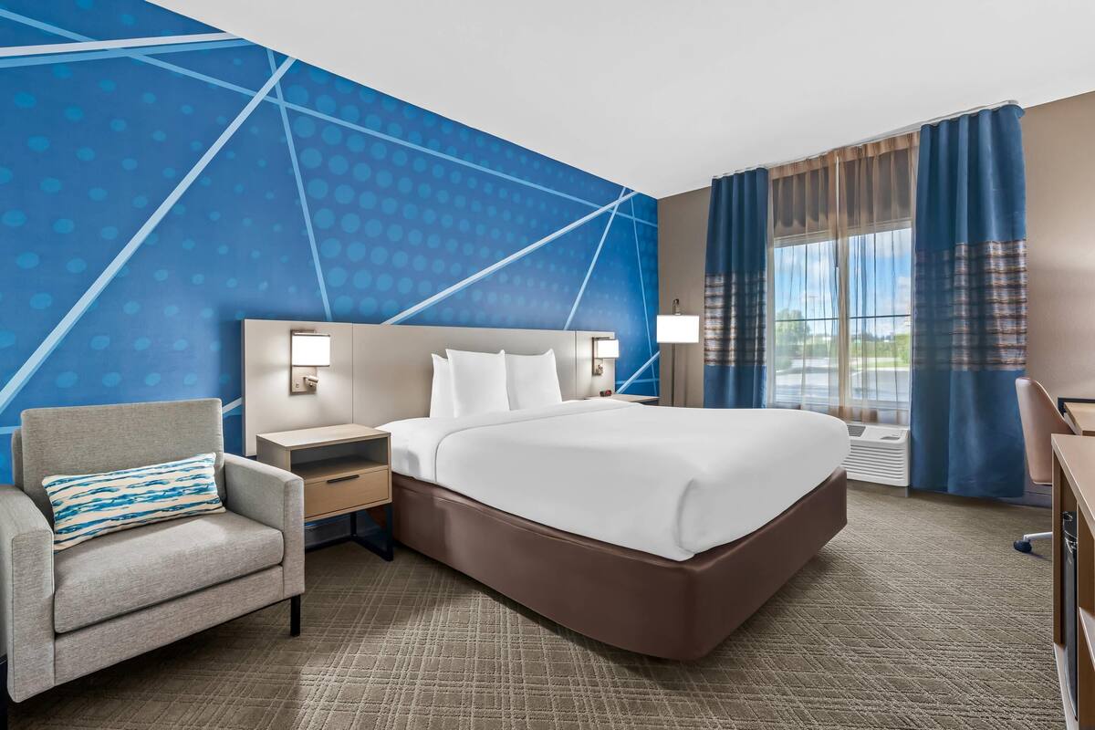 Habitación de hotel con cama, una silla, mesita de noche, lámpara y ventana con cortinas.