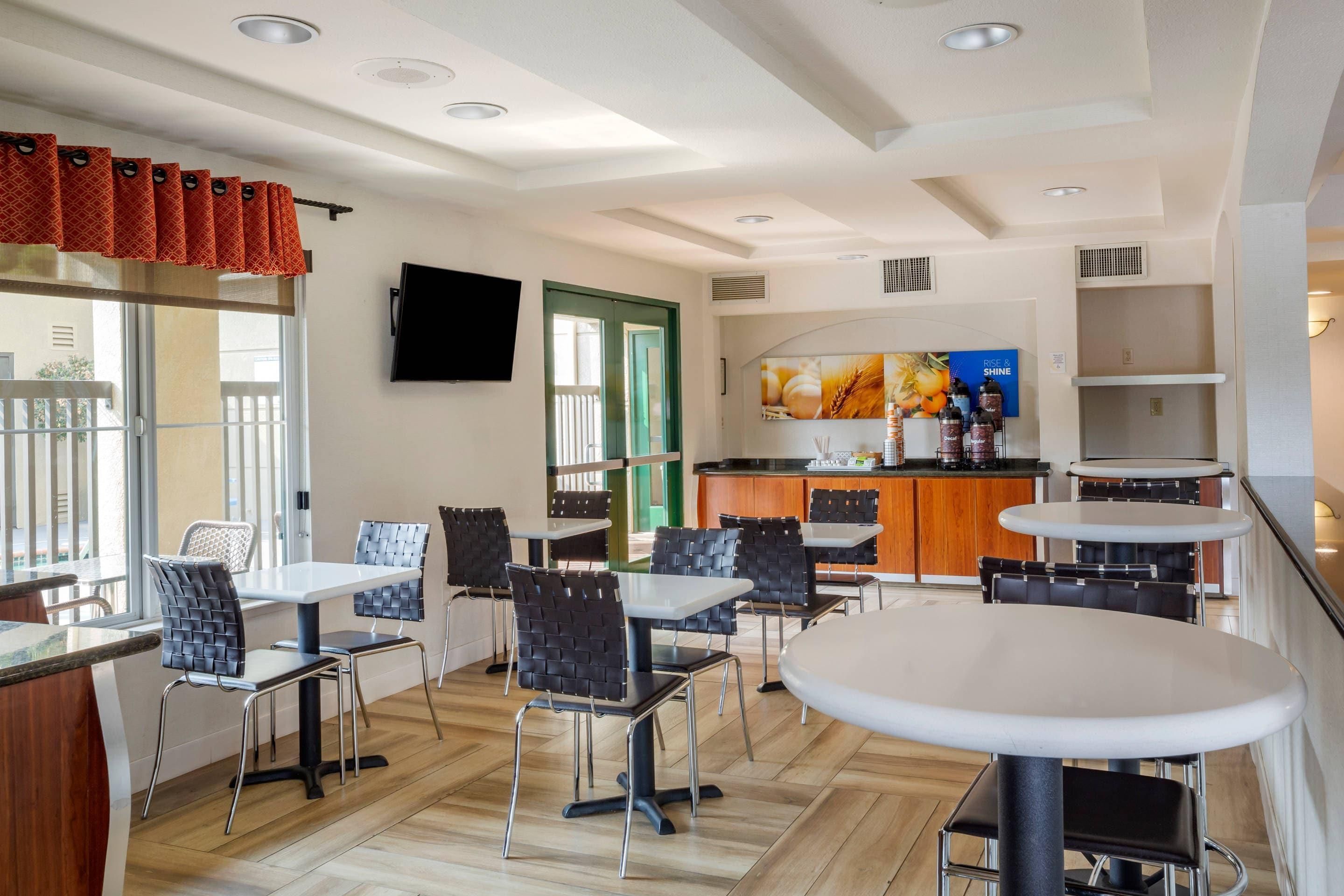 A modern dining area with tables and chairs, a kitchen area with a counter and appliances, and a television mounted on the wall.
