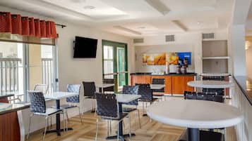 A modern dining area with tables and chairs, a kitchen area with a counter and appliances, and a television mounted on the wall.