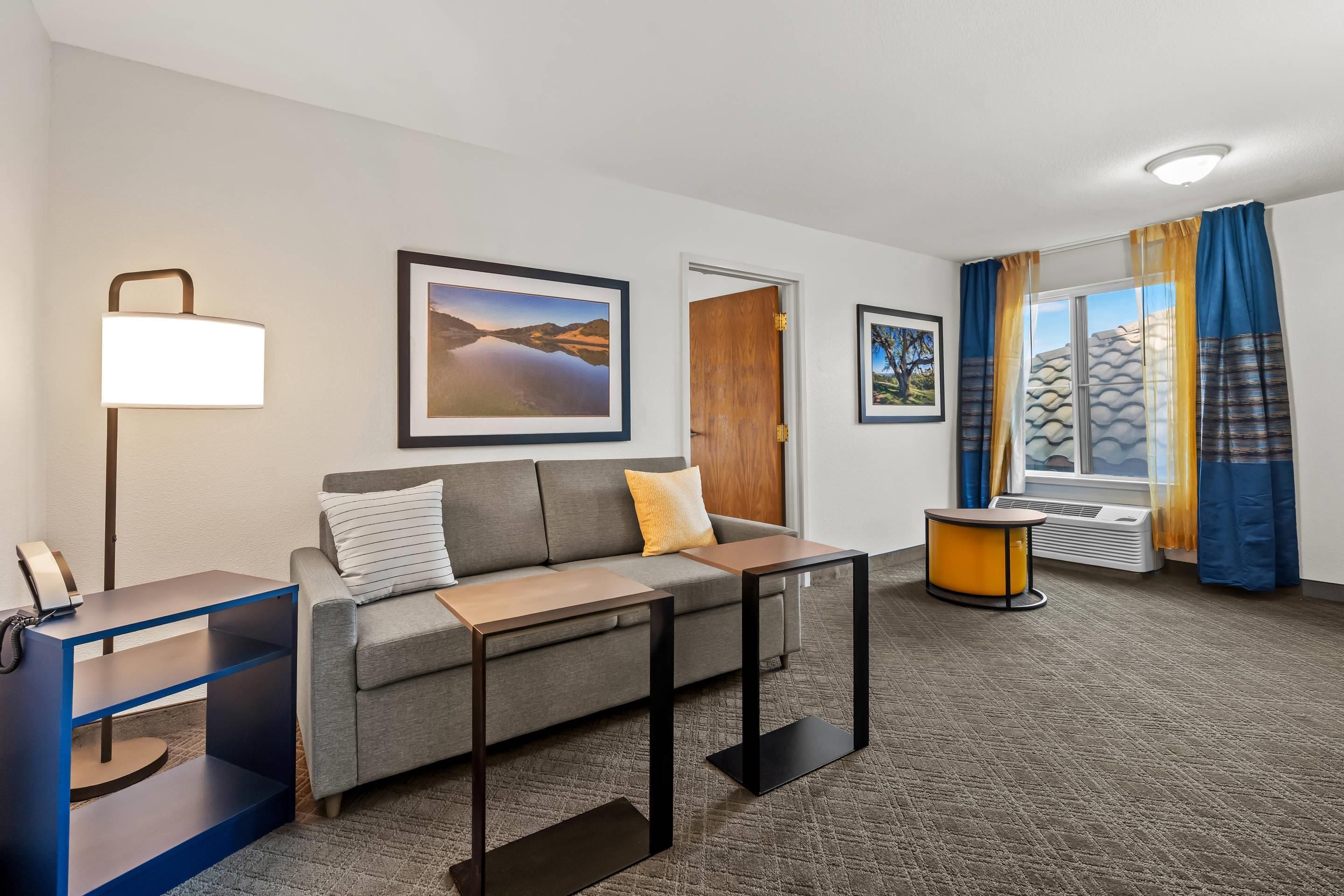 A hotel room with a sofa, a desk, a lamp, framed pictures on the wall, a window with curtains, and an air conditioning unit.
