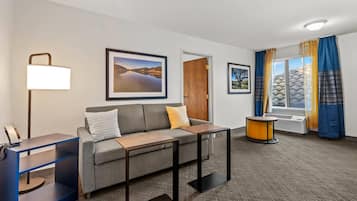A hotel room with a sofa, a desk, a lamp, framed pictures on the wall, a window with curtains, and an air conditioning unit.