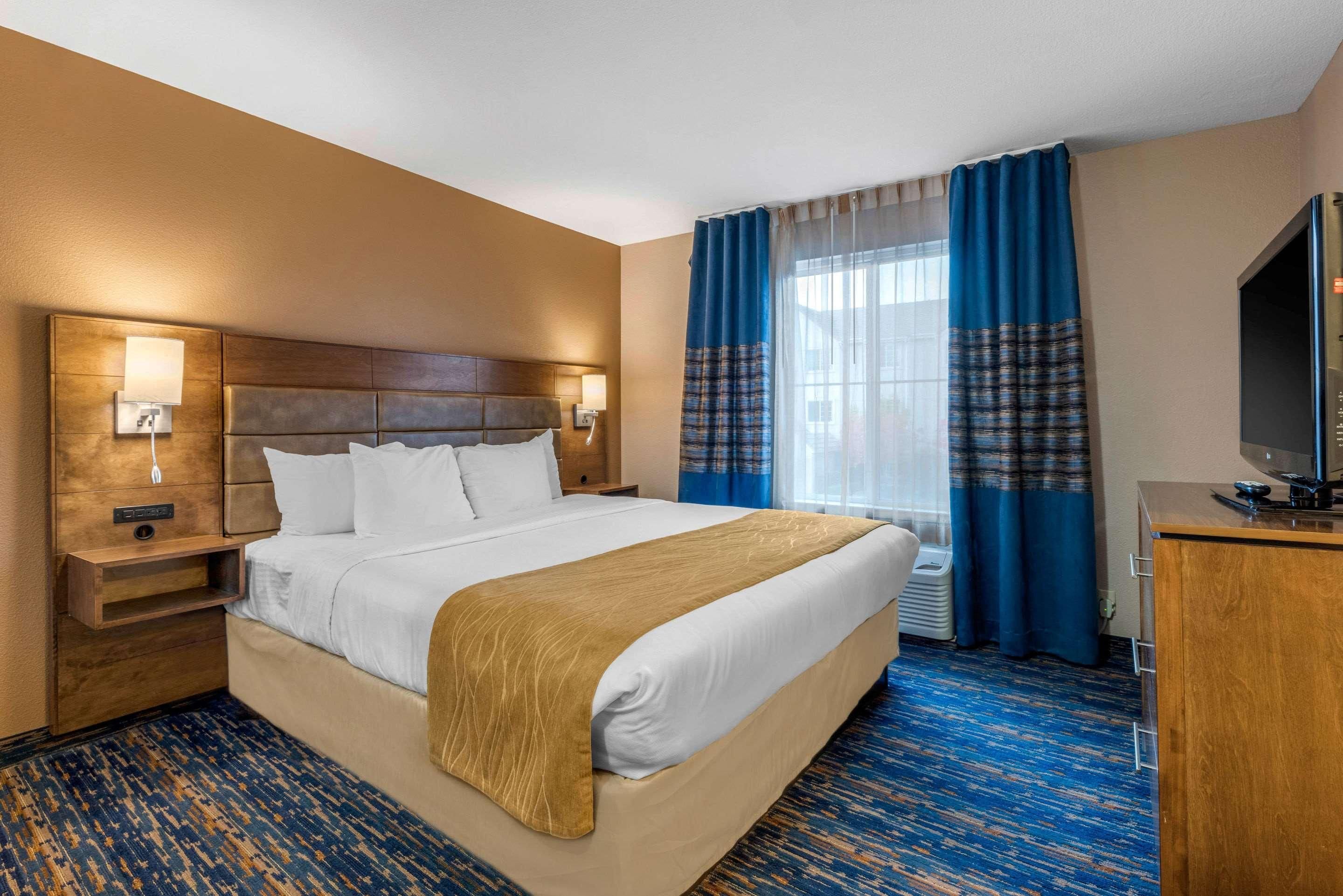 A hotel room with a large bed, a wooden headboard, a nightstand, a TV, and a window with curtains.