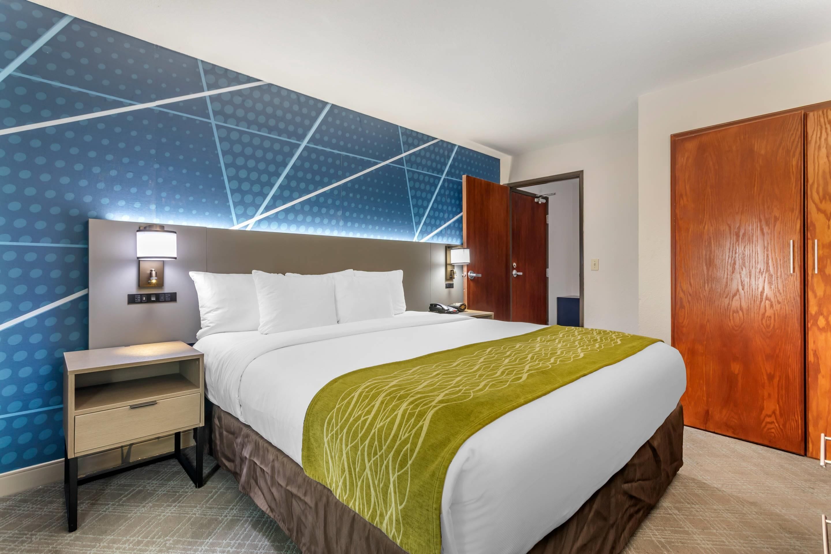 A hotel room with a large bed, a nightstand with a lamp, a wooden wardrobe, and a blue patterned wall.