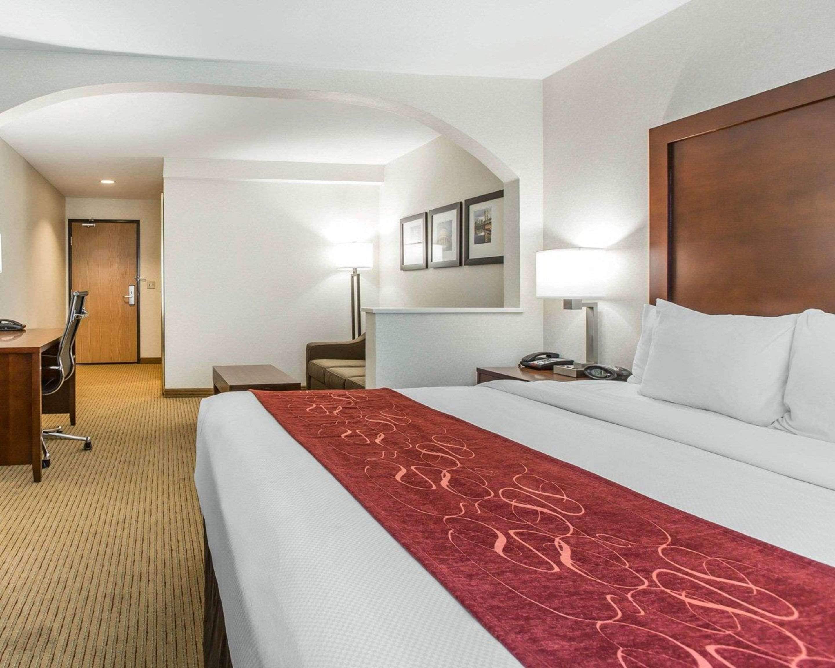 Una habitació d'hotel amb un llit gran, una taula de treball, una cadira, una làmpada i una porta que condueix a una altra habitació.
