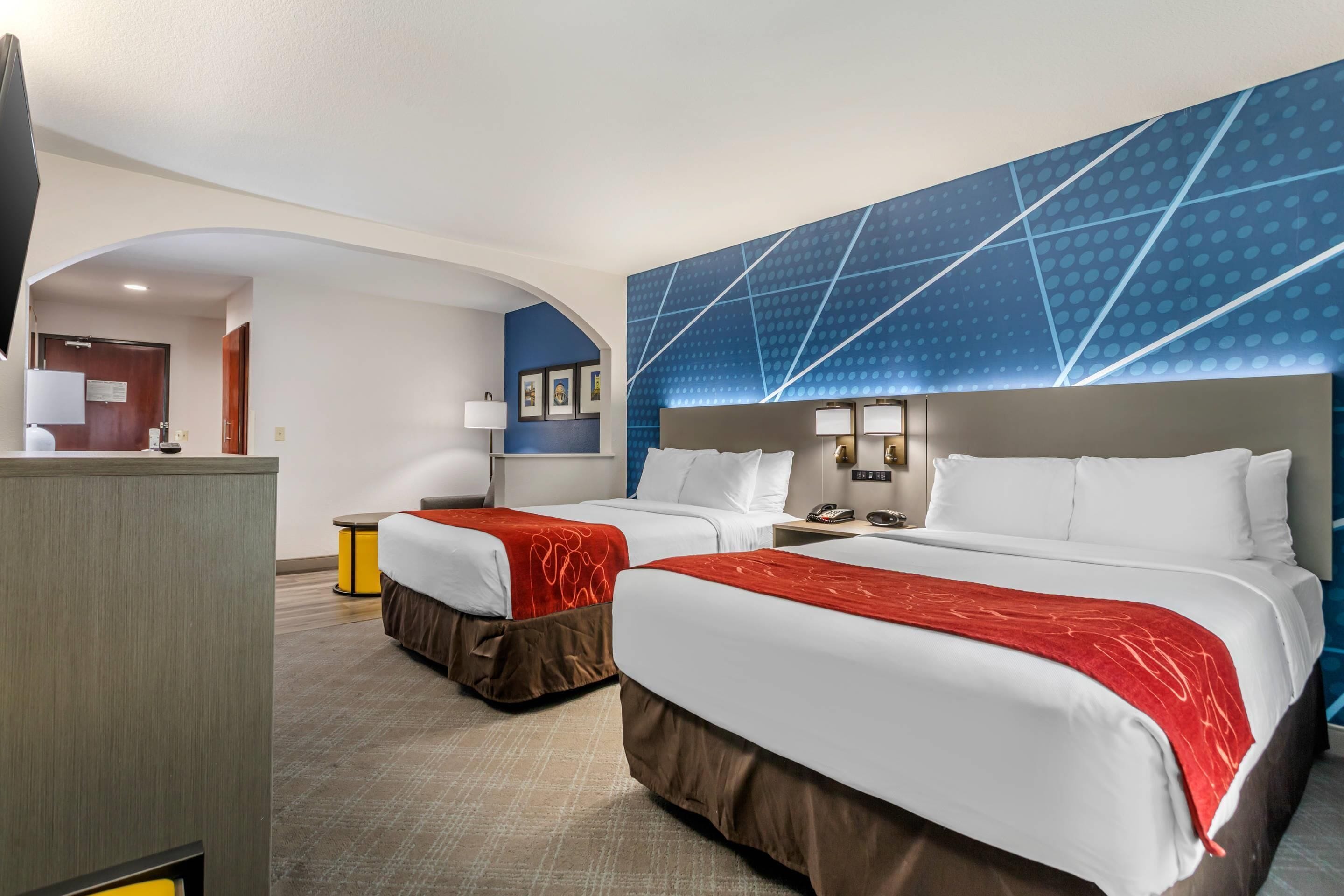 A hotel room with two beds, a TV, a desk, and a blue accent wall.