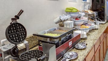 A buffet setup with a waffle maker, various food containers, and a refrigerator in the background.