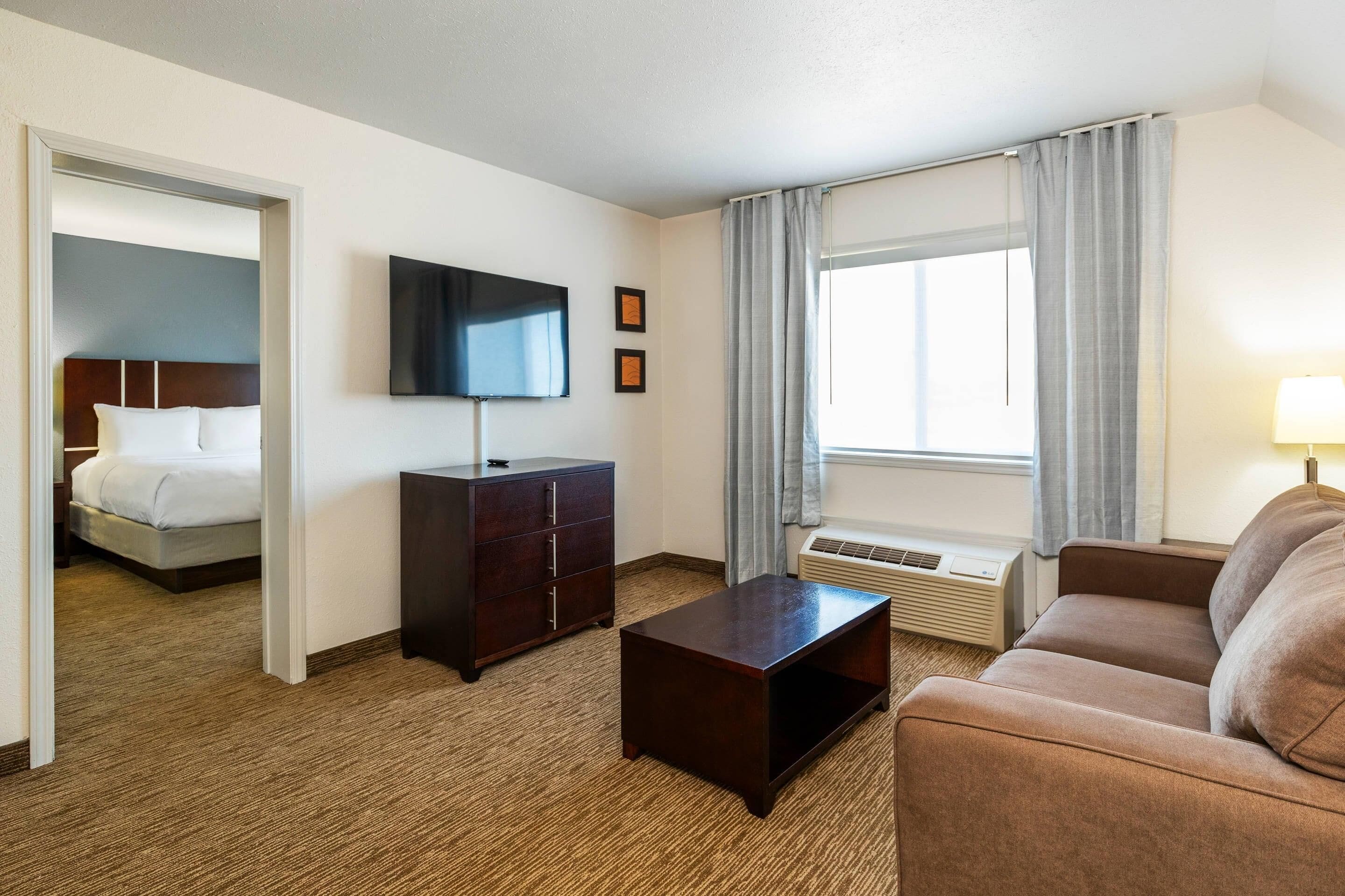 Quarto de hotel com cama, televisão, penteadeira, mesa de centro, sofá e janela com cortinas.