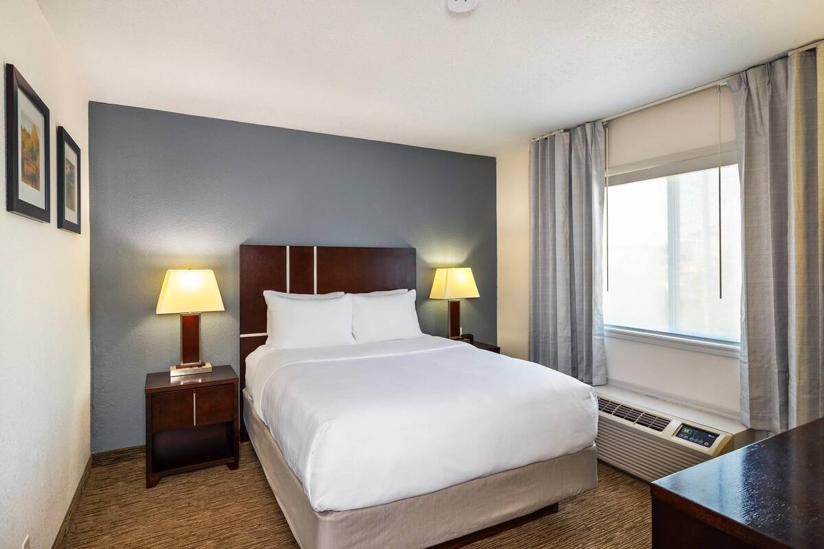 A hotel room with a large bed, two bedside lamps, a window with curtains, and a wall-mounted air conditioning unit.
