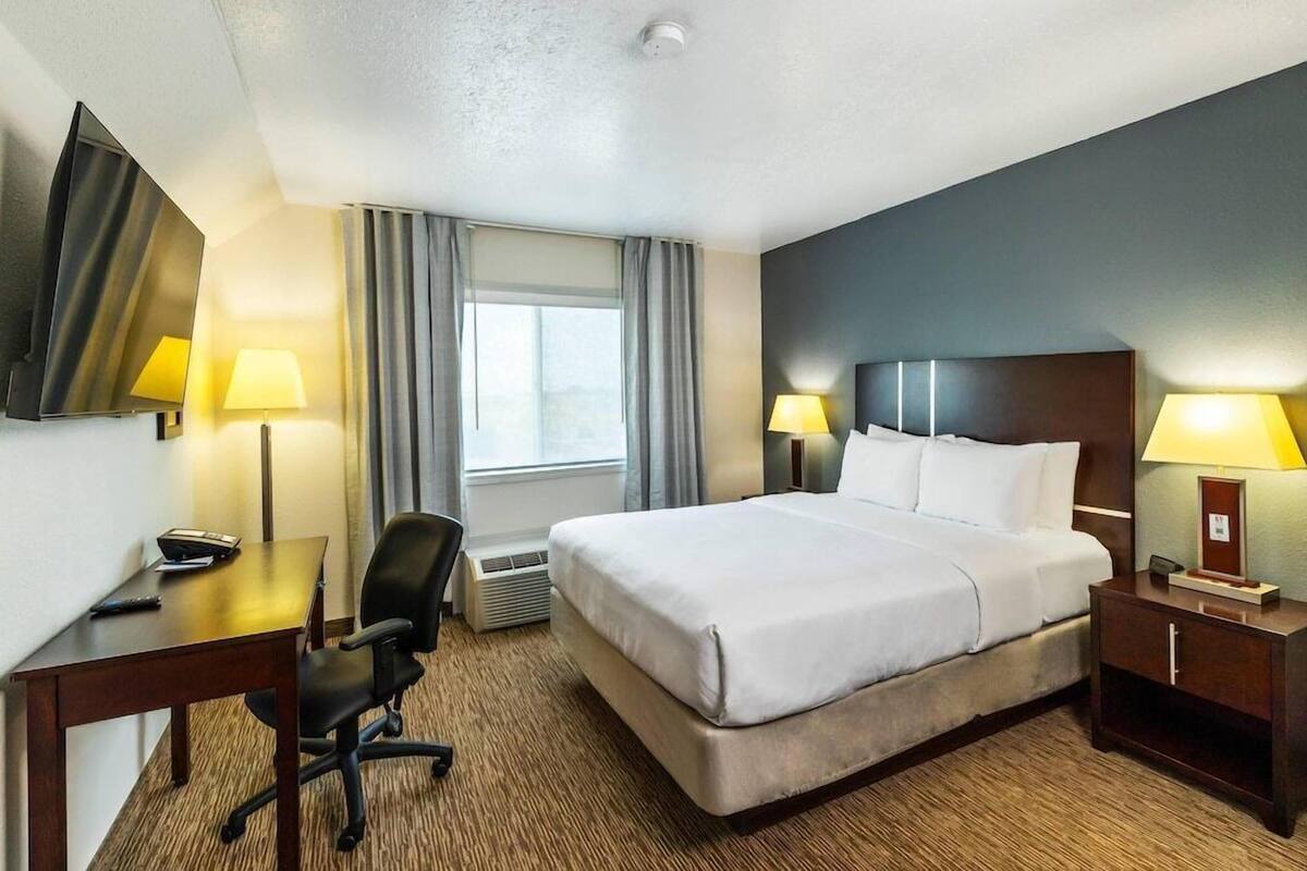 A hotel room with a bed, desk, chair, two bedside lamps, a TV, and a window with curtains.