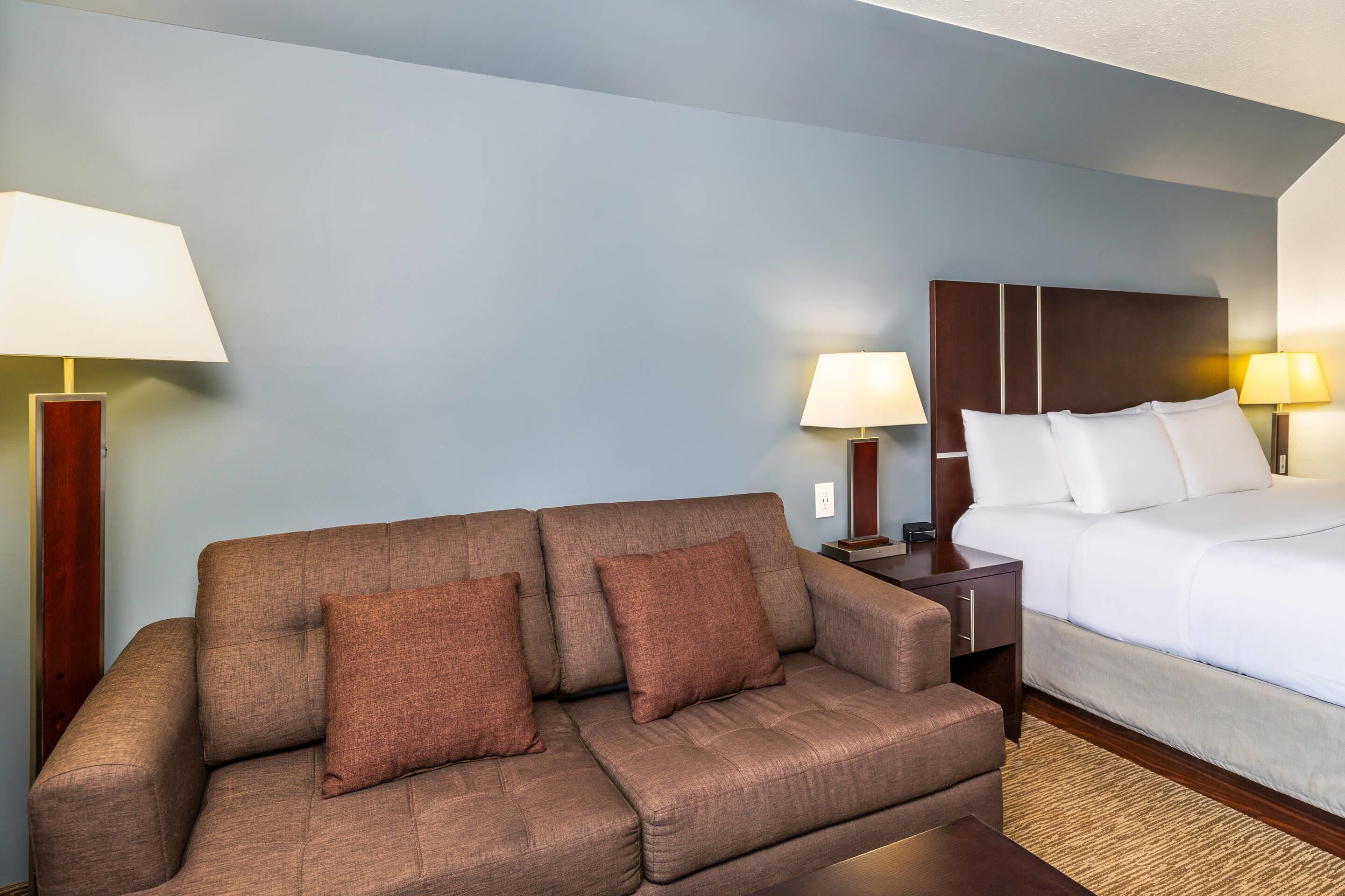Una habitación de hotel con un sofá marrón, una cama con ropa de cama blanca y dos lámparas de noche.