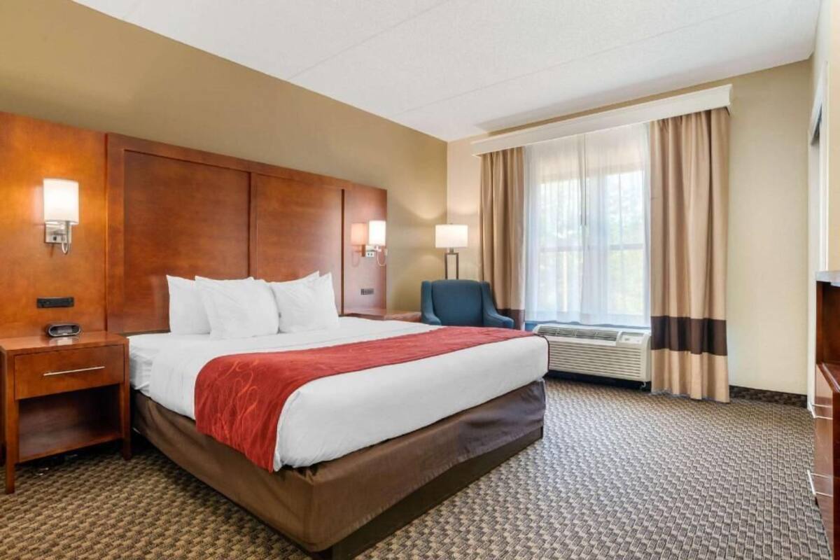 A hotel room with a large bed, bedside tables, a chair, a window with curtains, and a wall-mounted lamp.