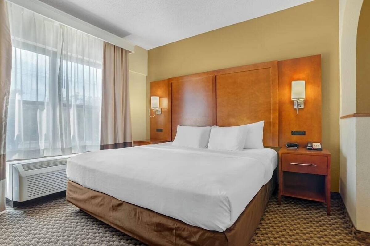 A hotel room with a large bed, two bedside tables, a wall-mounted lamp, and a window with sheer curtains.