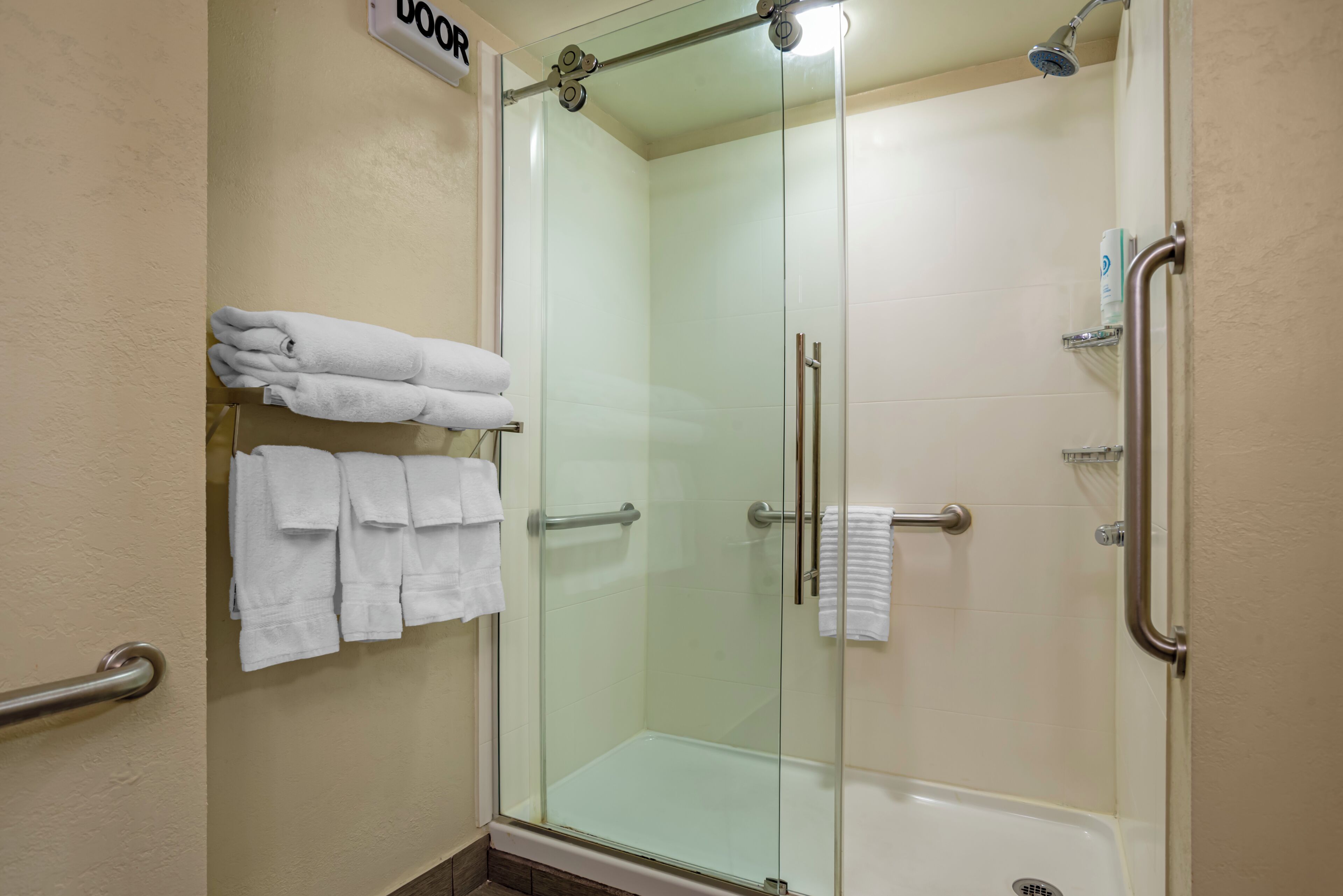 A glass-enclosed shower with a handrail, a towel rack with folded towels, and a sign indicating the door.