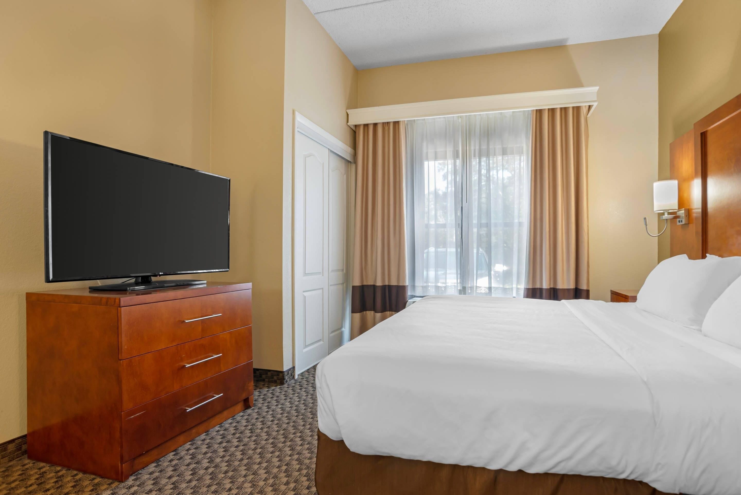 Una habitación de hotel con cama, televisor sobre una cómoda de madera, ventana con cortinas y puerta.