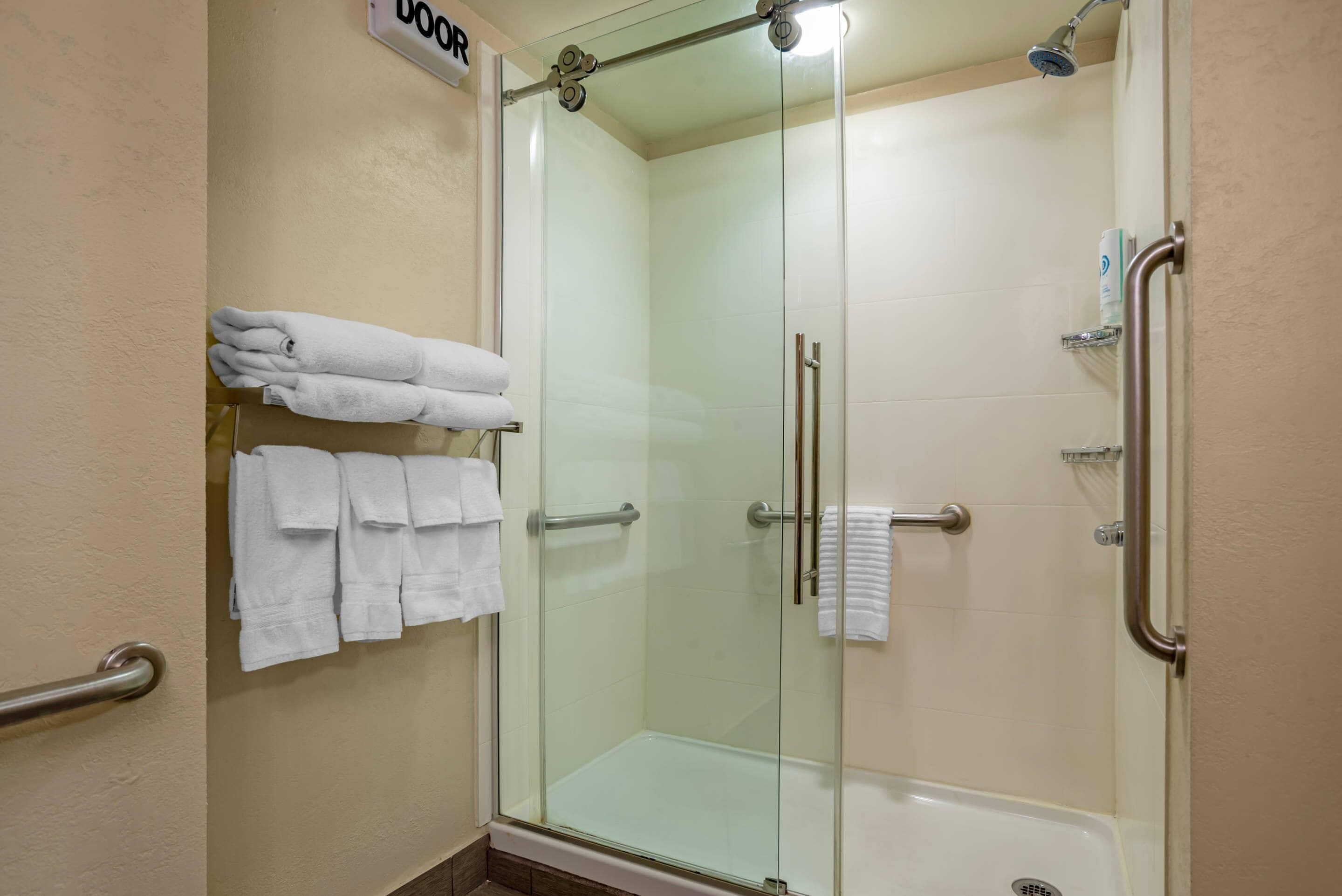 A glass-enclosed shower with a handrail, a towel rack with folded towels, and a sign above the door.