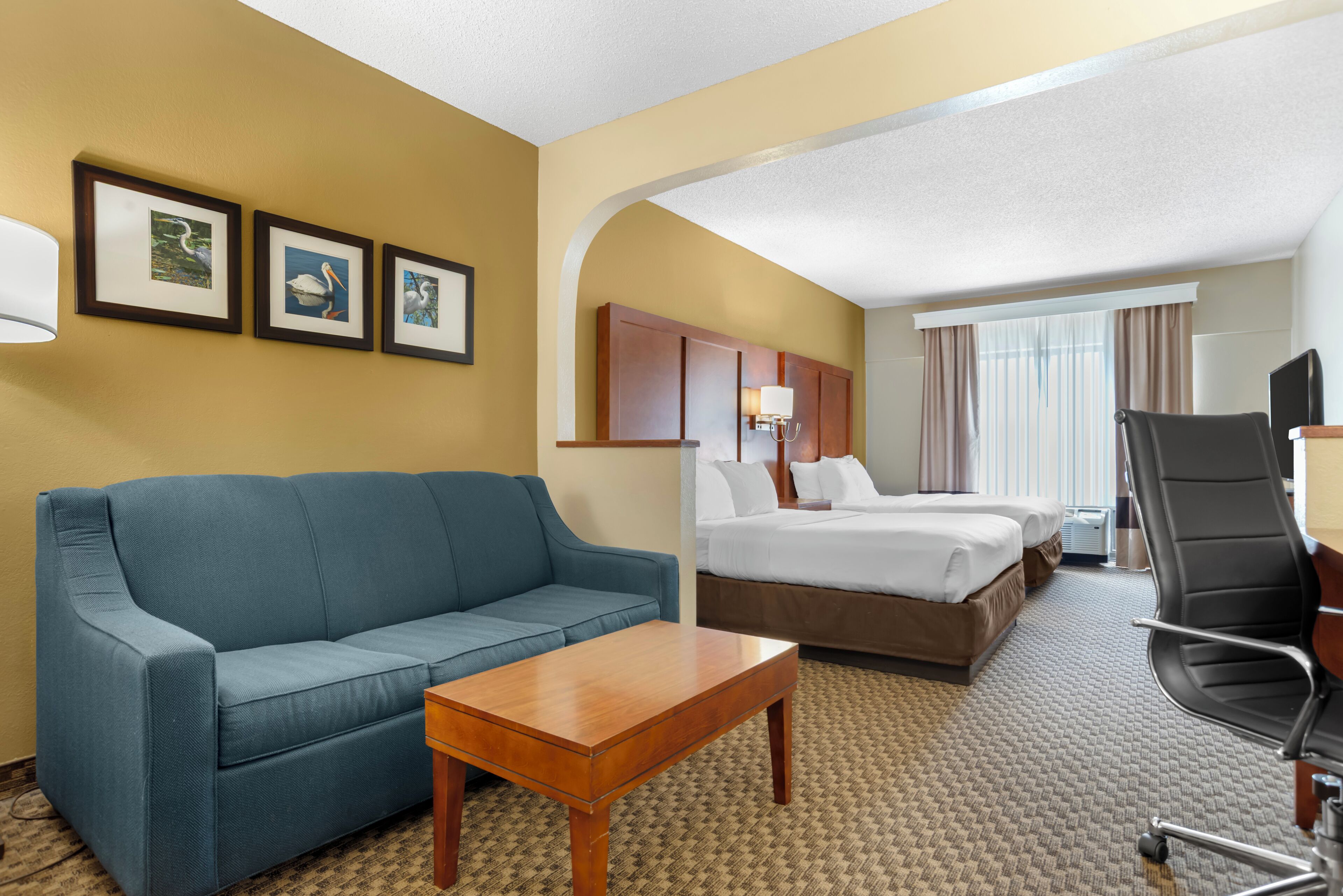 A hotel room with a blue sofa, a wooden coffee table, a bed with white linens, a black office chair, and framed pictures on the wall.