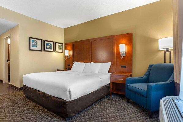 A hotel room with a large bed, a blue armchair, a wooden headboard, wall-mounted lamps, and framed pictures on the wall. - Comfort Suites Tallahassee Capitol - University Area (Tallahassee)