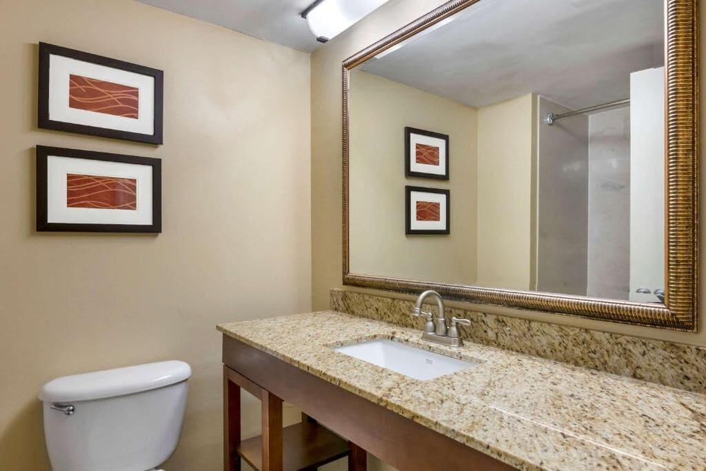 Un baño con inodoro, lavamanos y un espejo grande. Hay dos cuadros en la pared.