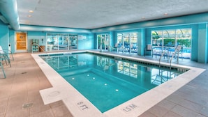 A swimming pool with clear water, surrounded by blue walls and a tiled floor. There are lounge chairs and a window with a view of greenery outside.