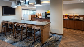 A modern hotel lobby with a large wooden reception desk, multiple bar stools, a coffee station, and a flat-screen TV.