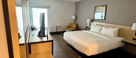 A modern hotel room with a large bed, a desk with a chair, a desk lamp, and a small table.