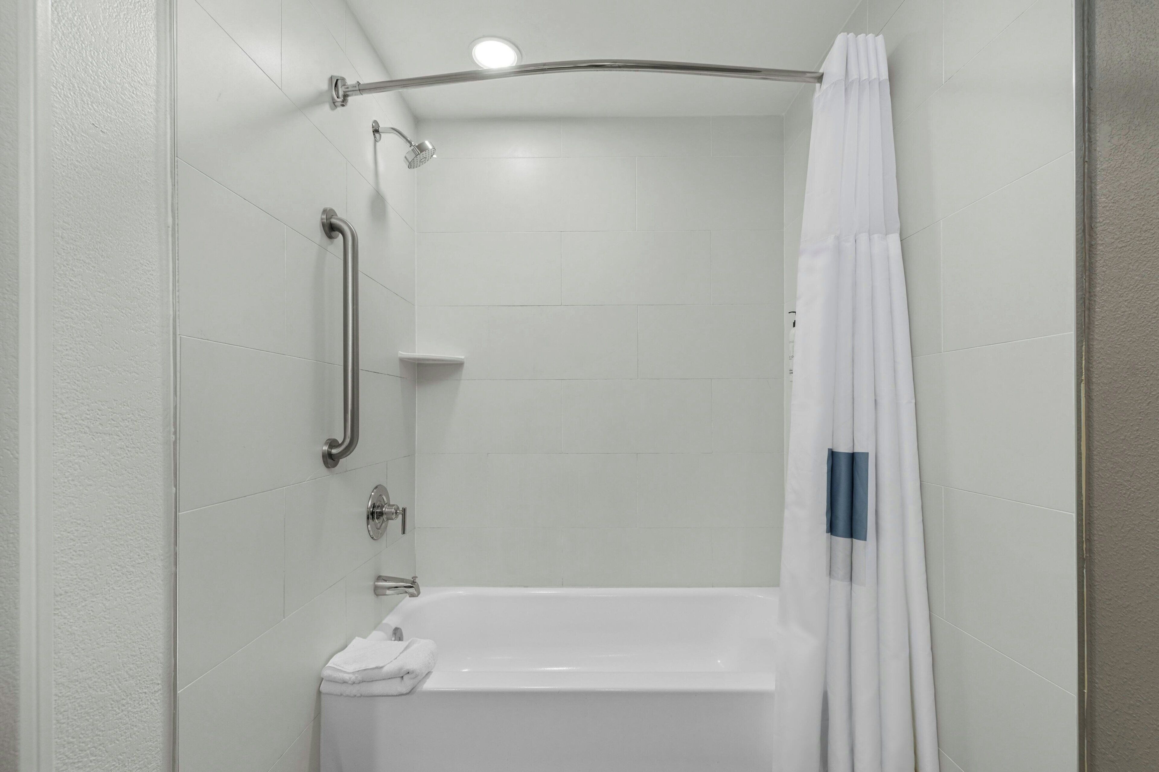 A modern shower area with a white bathtub, a handheld showerhead, a grab bar, and a white shower curtain with a blue stripe.