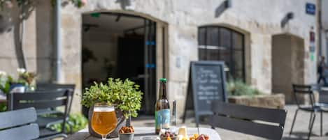 Une table dressée avec un plateau de fromages, une bouteille et un verre de bière, dans un espace de restauration en plein air aux murs de pierre et à l’ambiance rustique.