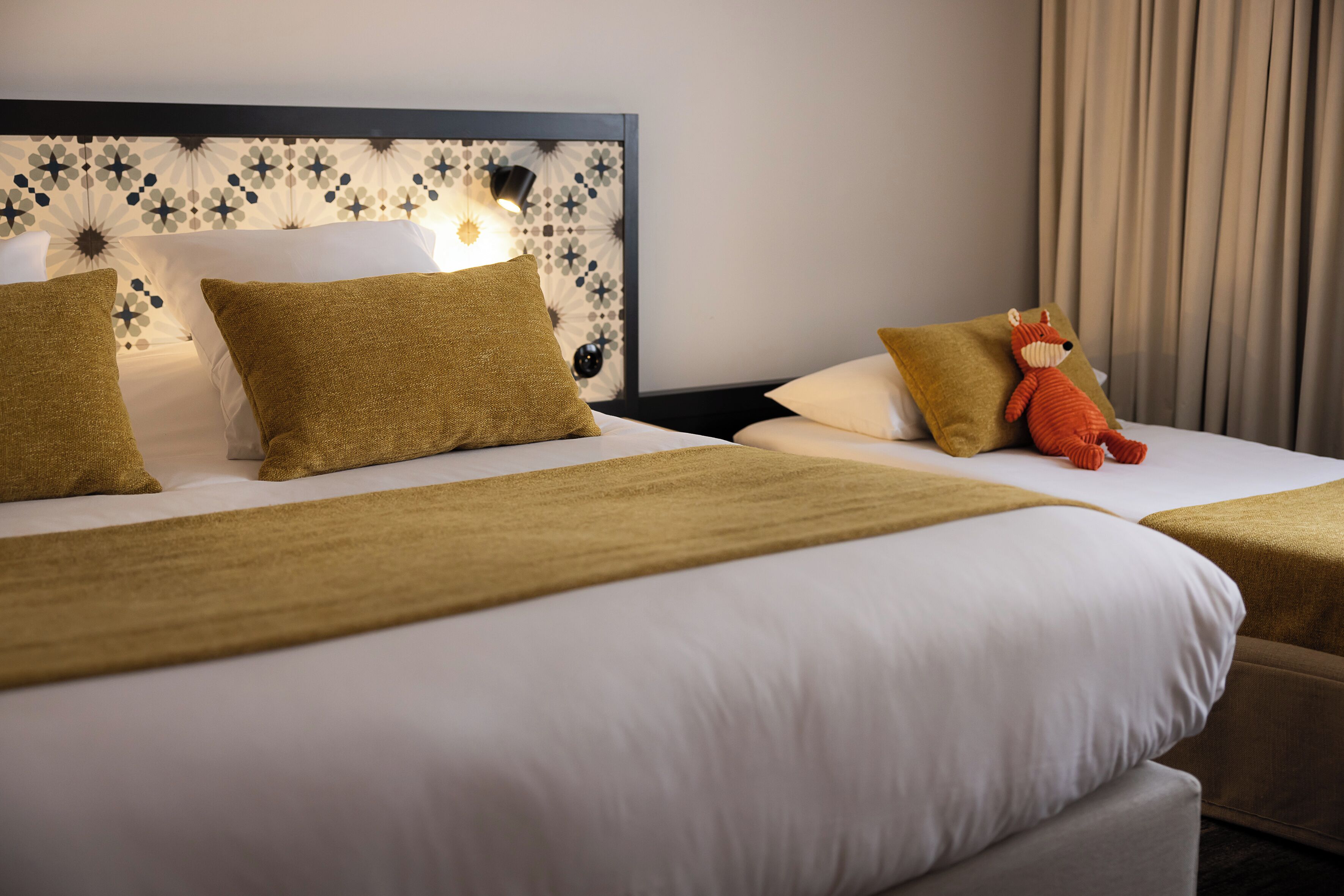 Une chambre d’hôtel avec deux lits, chacun doté d’une tête de lit ornée d’un motif géométrique. Un doudou est posé sur l’un des lits, et la chambre est décorée dans des tons neutres.