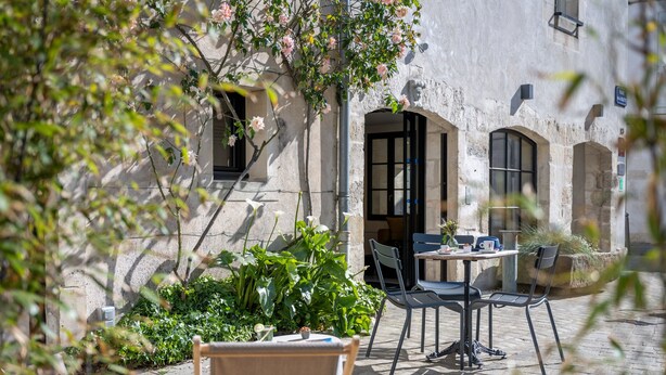 An outdoor seating area with a table and chairs, surrounded by greenery and a building with stone walls and arched doorways. - Hotel Saint Nicolas (La Rochelle)