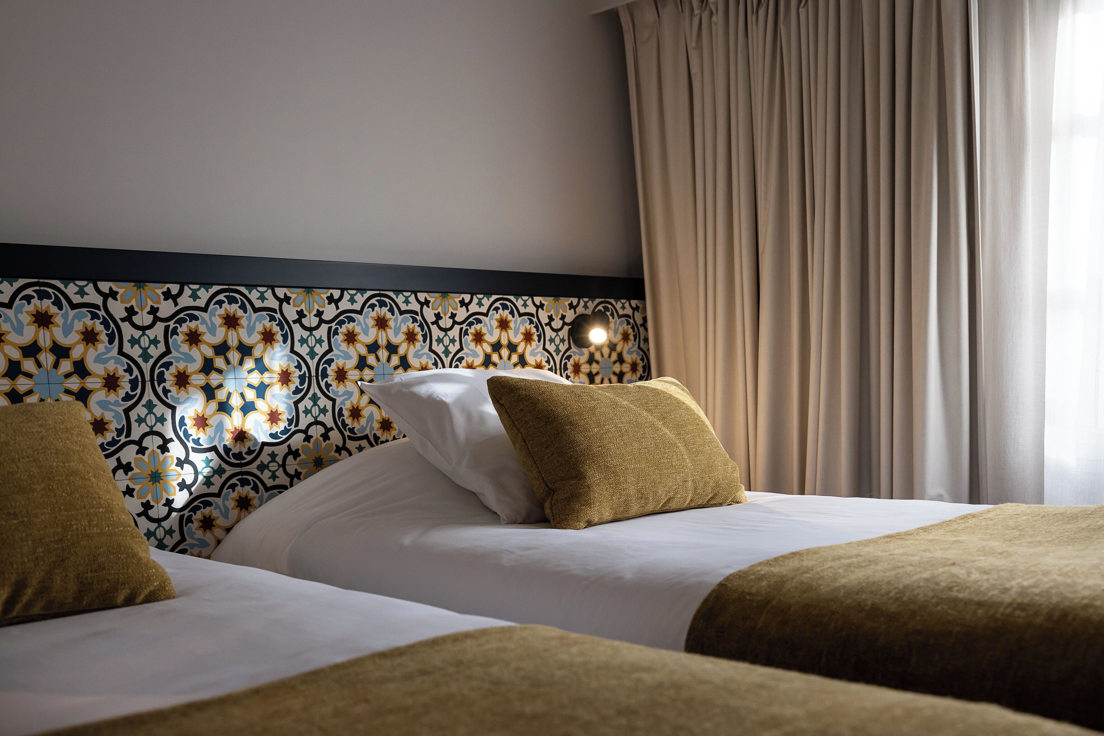 Une chambre d’hôtel avec une tête de lit à motifs, deux lits avec du linge de lit blanc et un rideau beige.