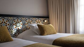 Une chambre d’hôtel avec une tête de lit à motifs, deux lits avec du linge de lit blanc et un rideau beige.