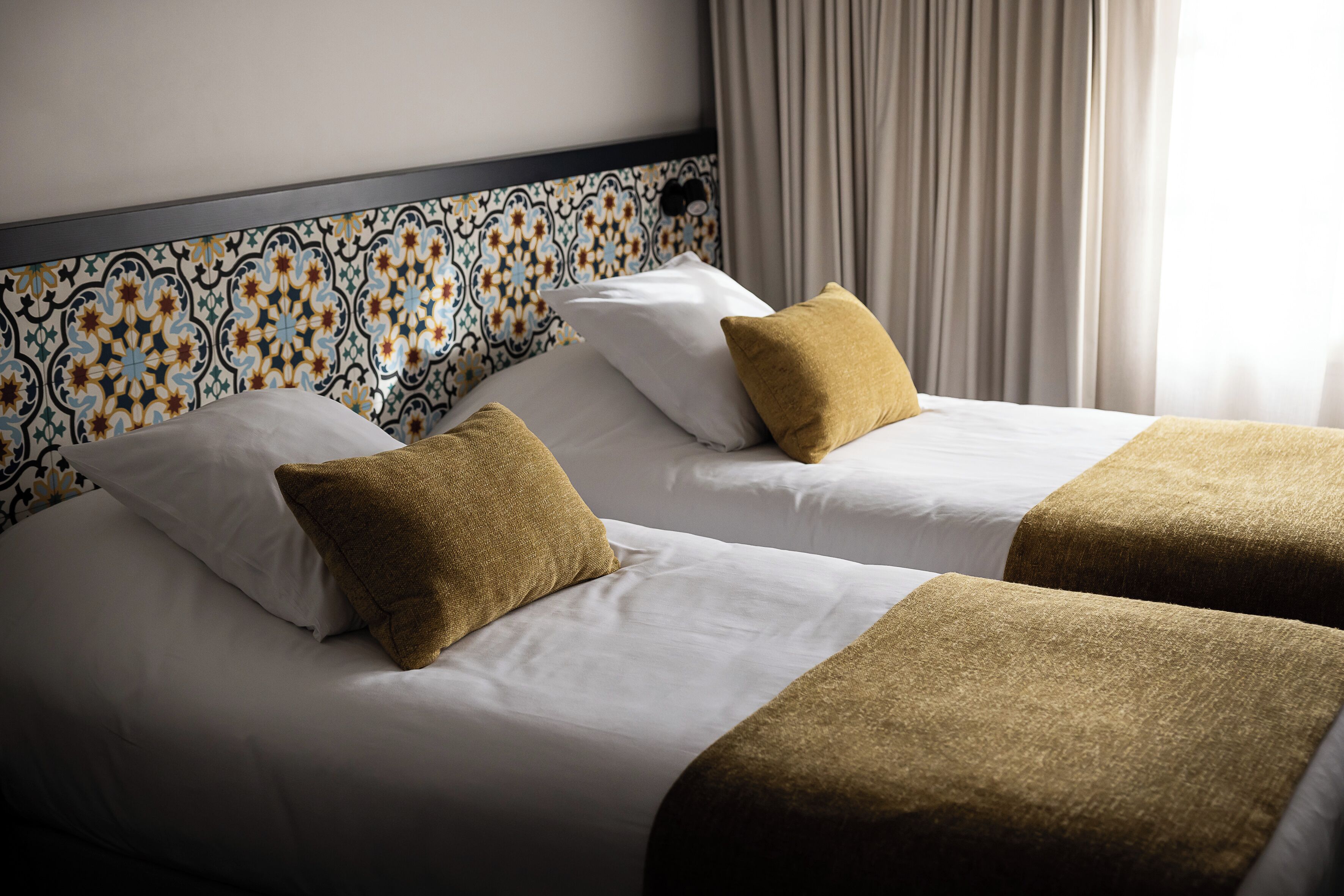 Two beds with white linens and brown pillows, a patterned headboard, and a window with curtains.