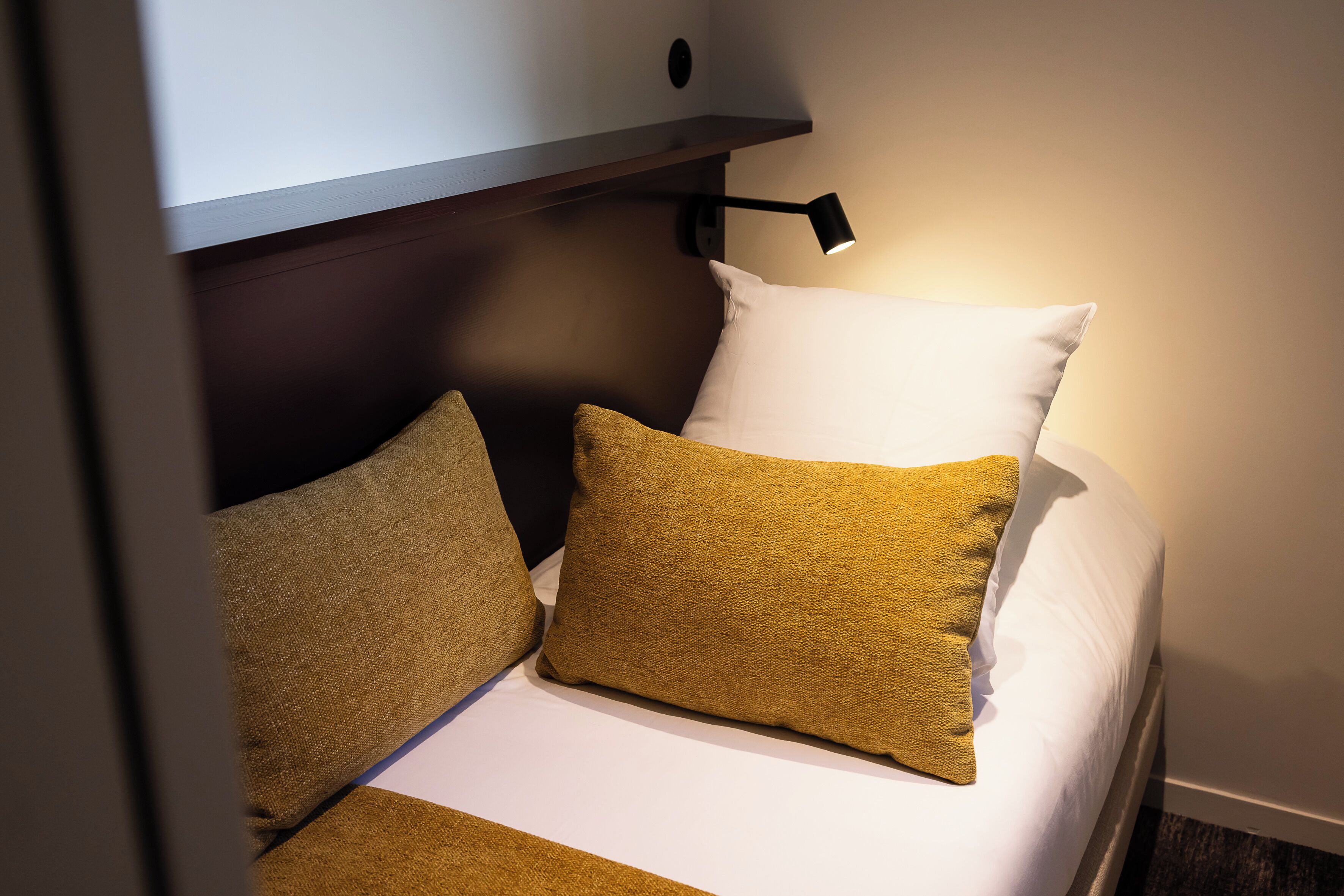 A neatly made bed with white and mustard-colored pillows, a black wall-mounted lamp, and a wooden headboard.