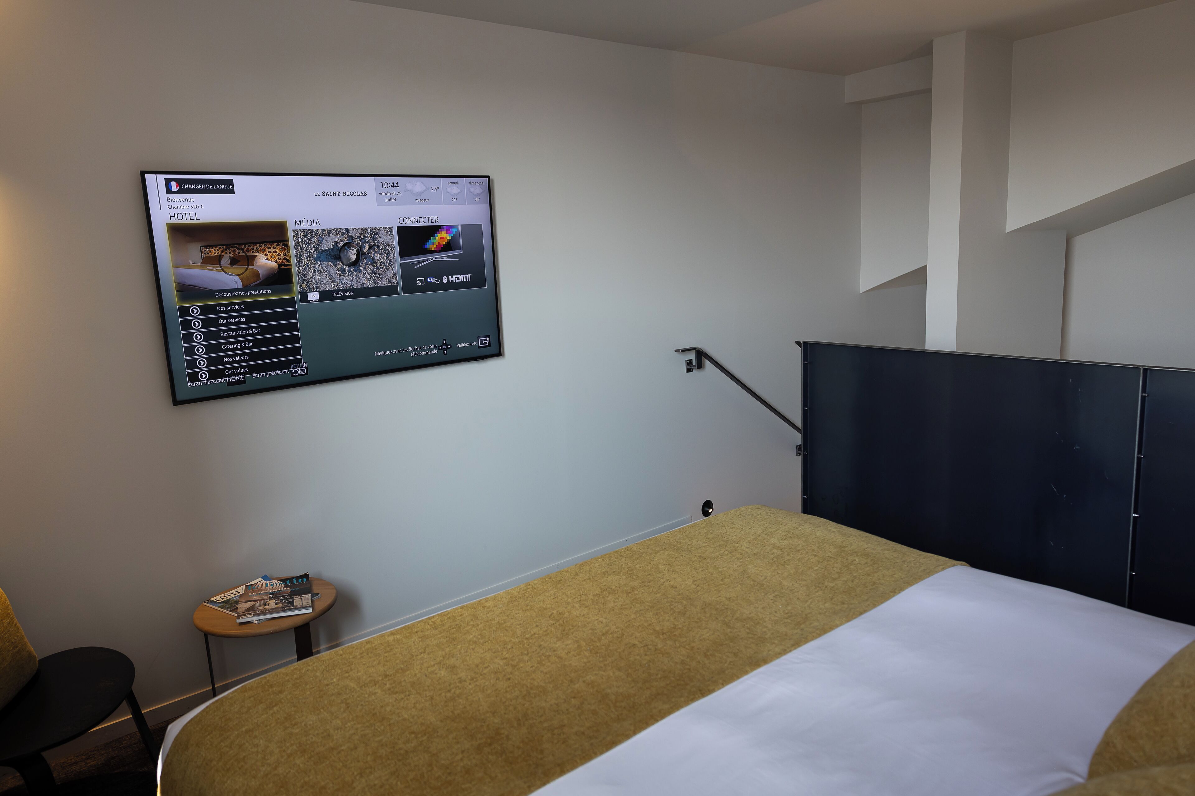 A hotel room with a bed, a small table with a magazine, and a TV displaying hotel information.