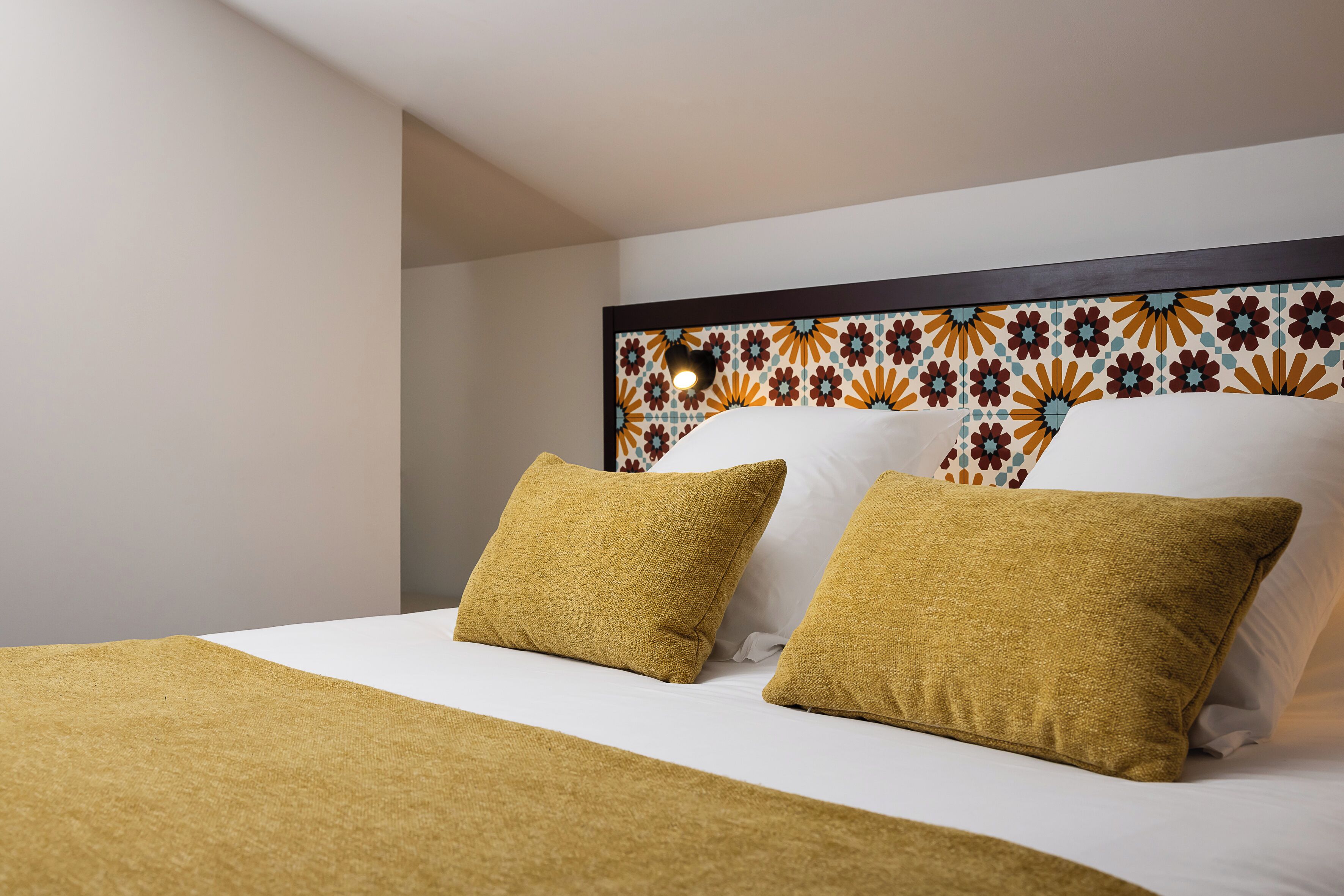 Una cama bien tendida con sábana blanca y dos almohadas color mostaza, cabecera estampada y pared lisa.