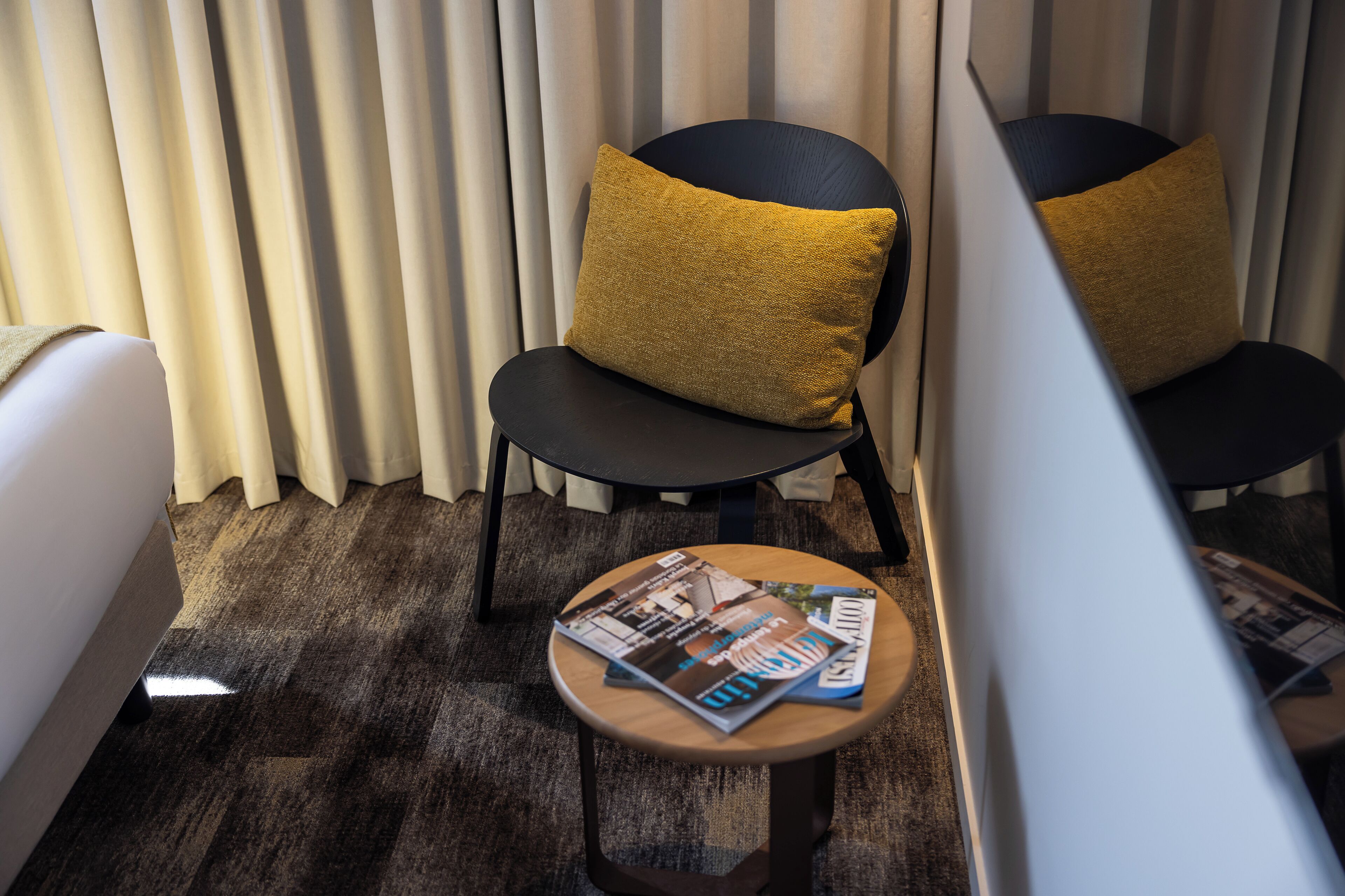 Une chambre d’hôtel avec un lit, deux chaises noires dotées de coussins de couleur moutarde, une petite table en bois avec des magazines et une fenêtre avec des stores verticaux.