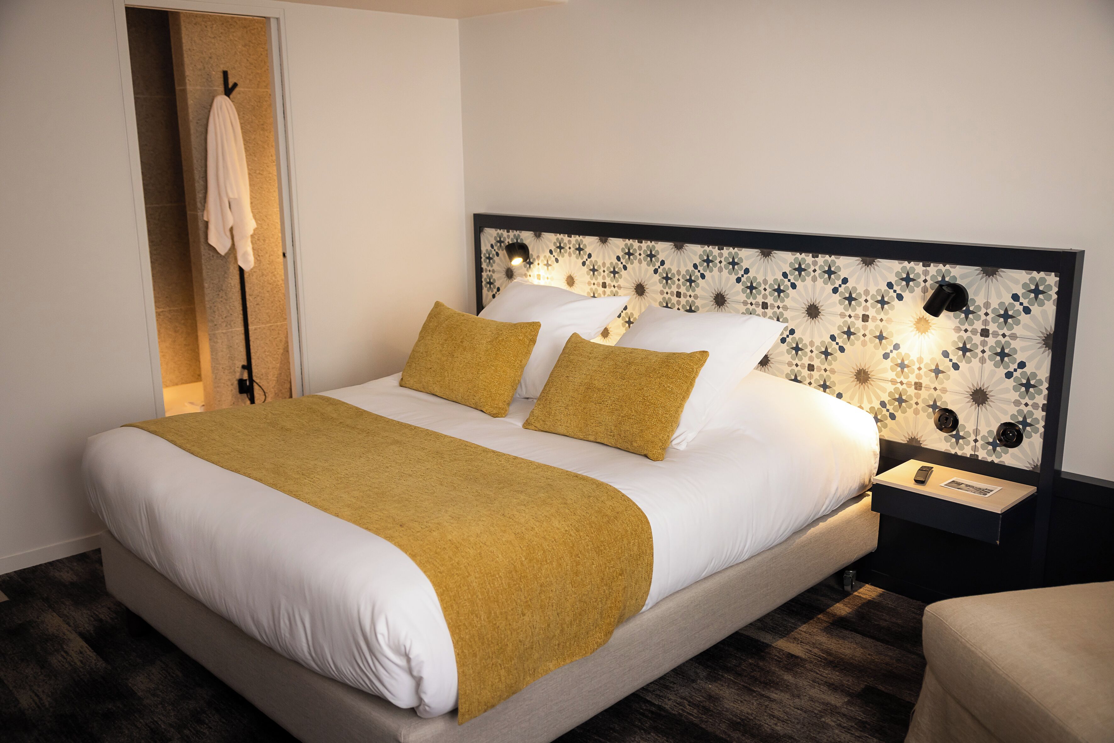 A neatly made bed with a white and mustard-colored bedspread, a headboard with a floral pattern, and a bedside table with a lamp.