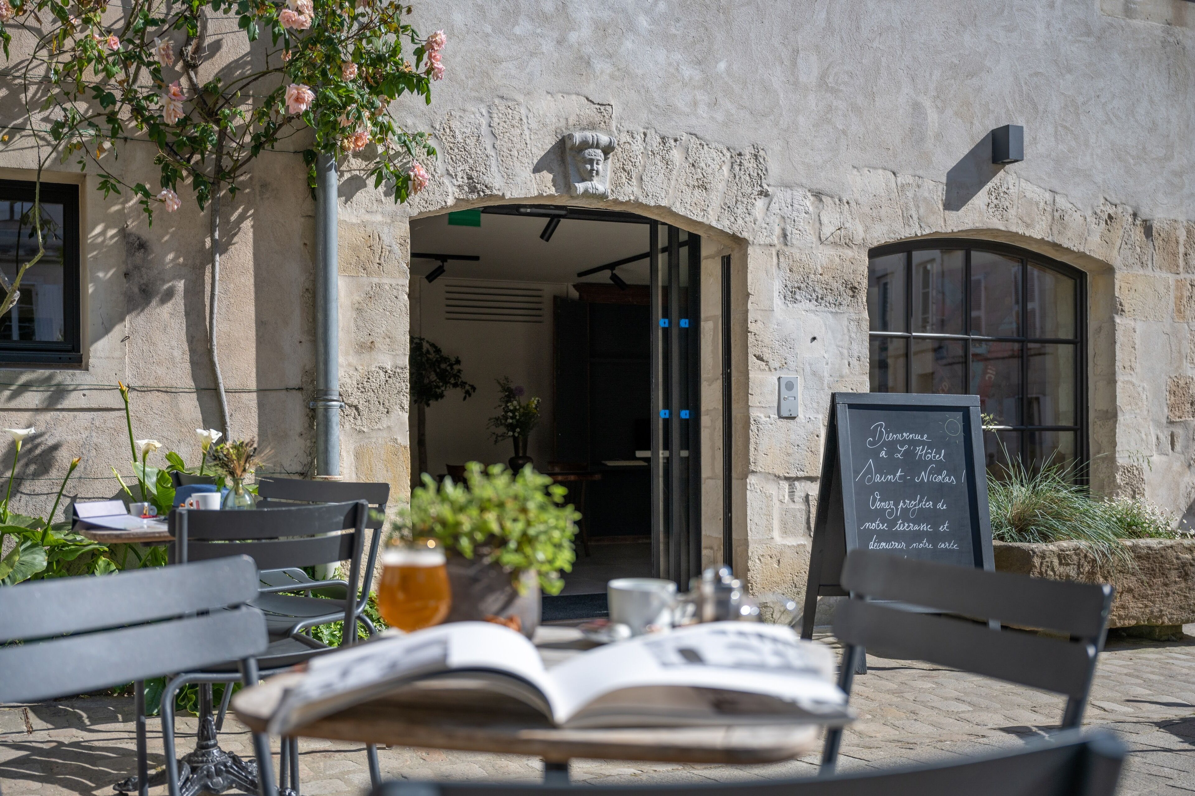 Un espace extérieur aménagé avec une table, des chaises et un banc. On y trouve un panneau noir avec du texte, une plante en pot et un mur en pierre.