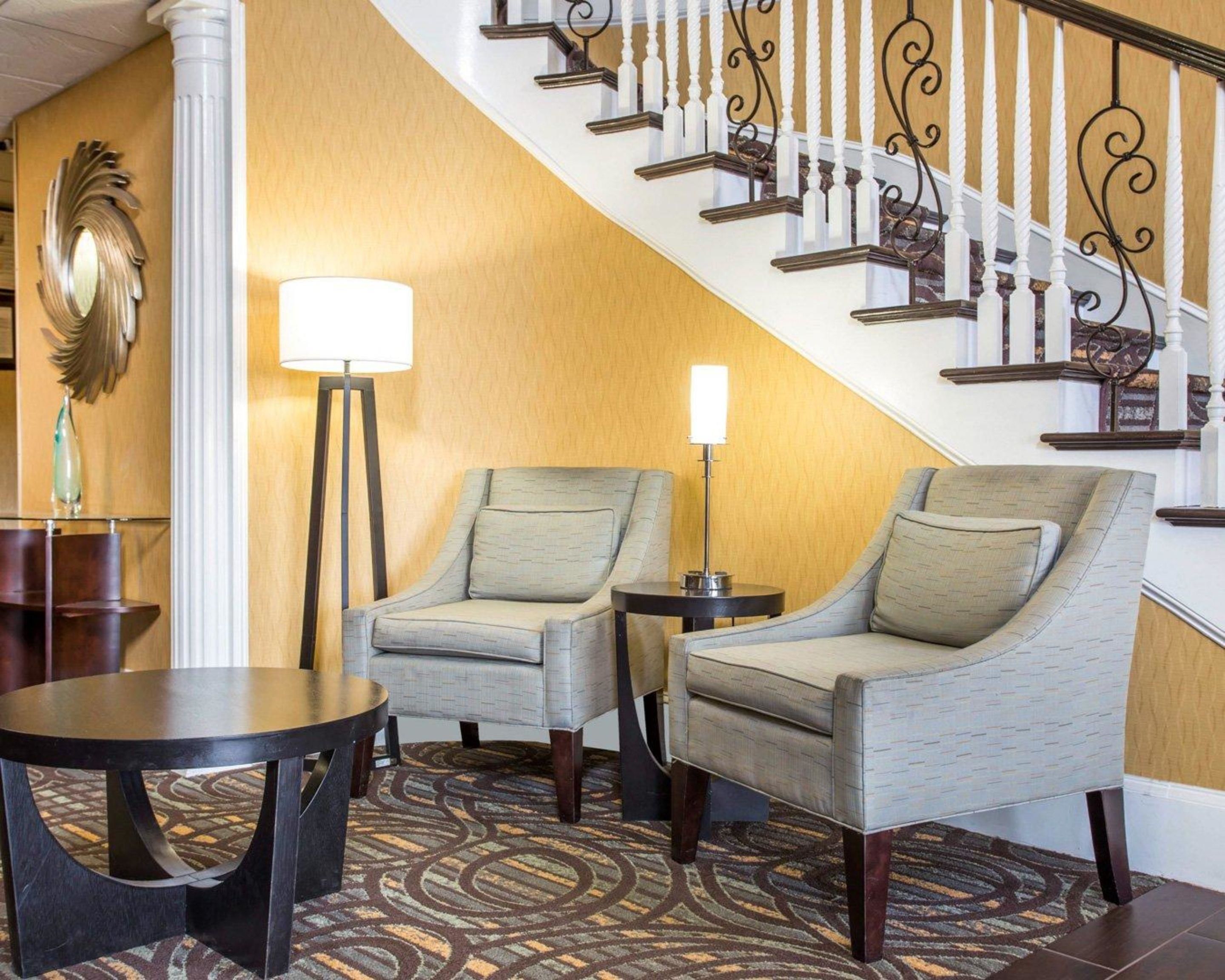 Un vestíbulo de hotel con una escalera, dos sillones, una mesa redonda y una lámpara de pie.