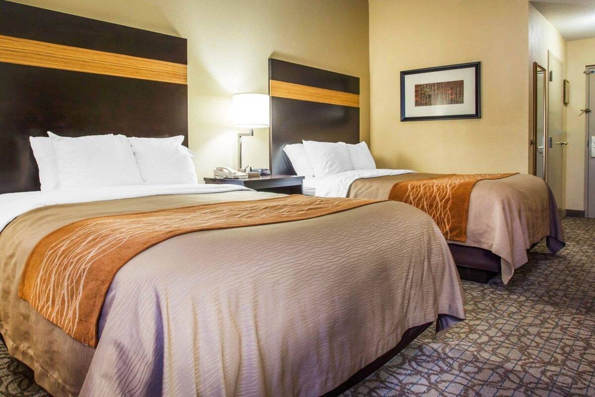 A hotel room with two beds, a nightstand with a lamp, a framed picture on the wall, and a door leading to another room.
