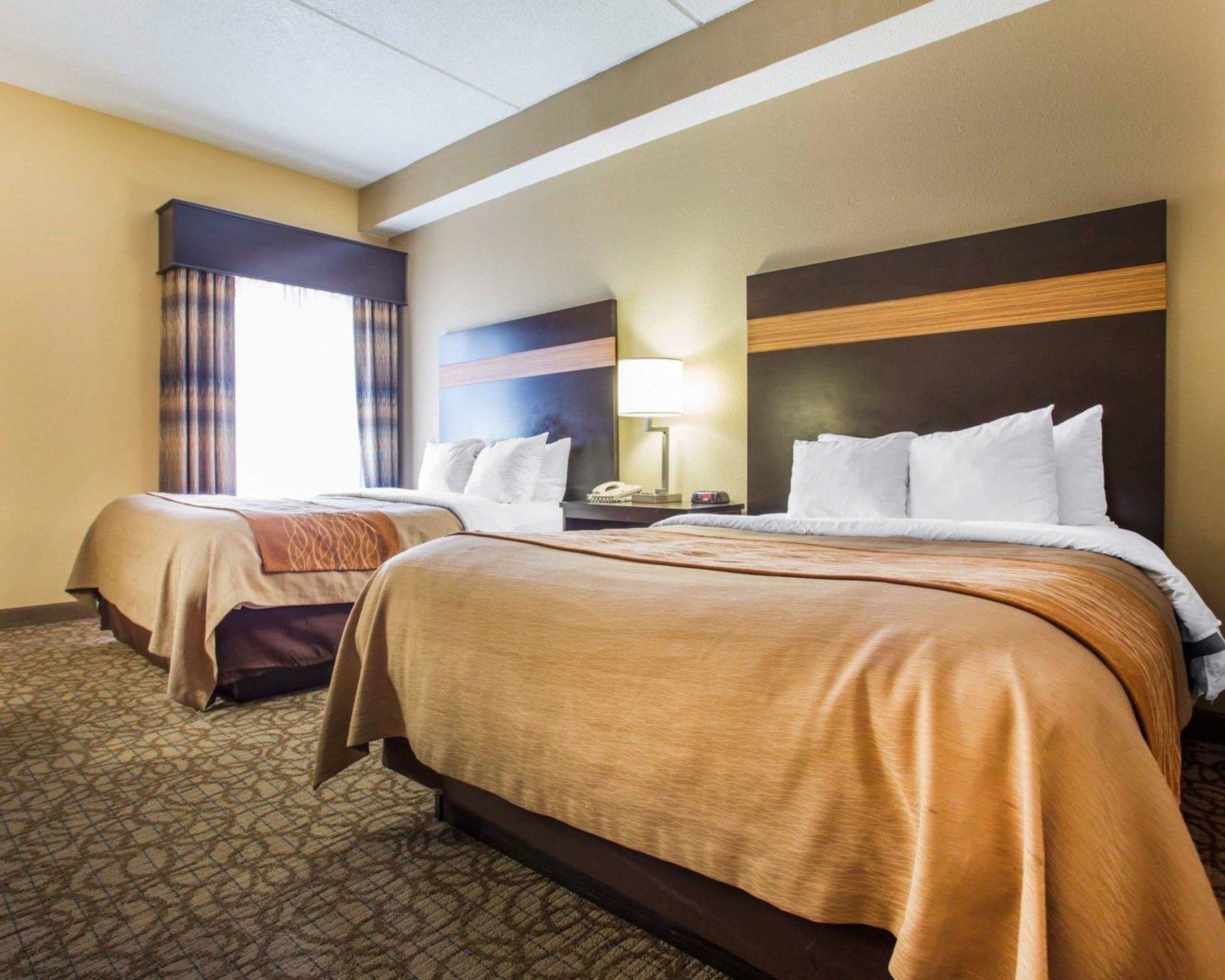 A hotel room with two beds, a nightstand, a lamp, and a window with curtains.
