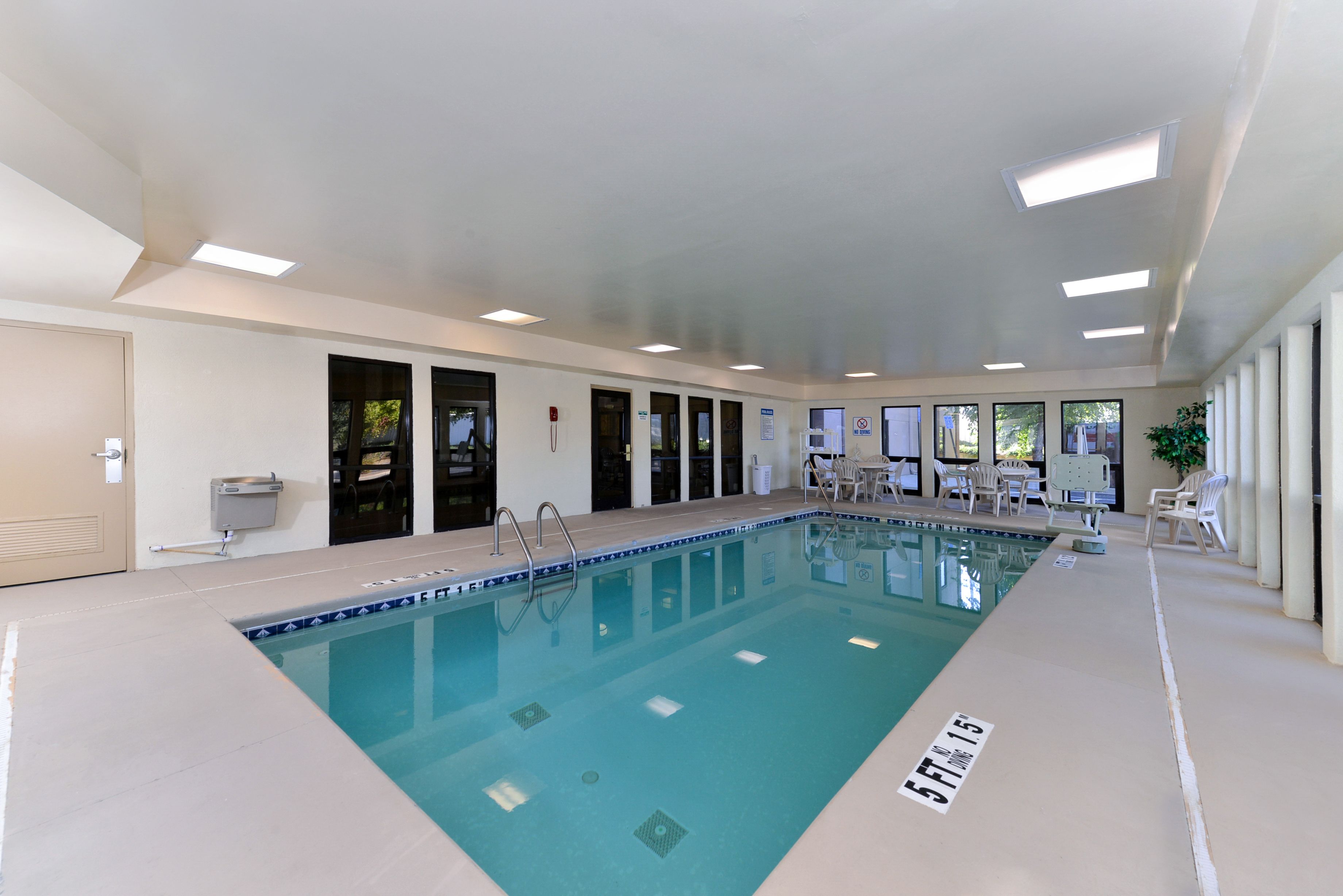 Uma piscina interior com água cristalina, rodeada por paredes e portas brancas, com luzes no teto e algumas cadeiras perto da piscina.