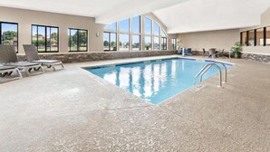 A large indoor swimming pool with clear views of the outdoors through large windows, a lounge area with chairs, and a stone wall surrounding the pool. - Comfort Suites near Robins Air Force Base (Warner Robins)