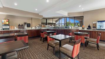 A hotel restaurant with multiple tables and chairs, a coffee machine, and a large window offering a view.