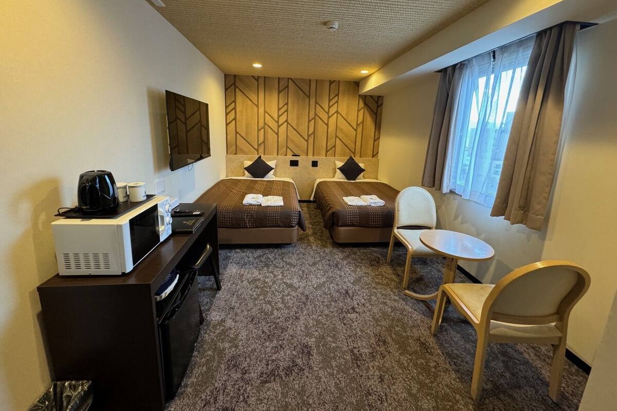 A hotel room with a bed, two bedside tables, a desk with a computer, a chair, and a window with curtains.