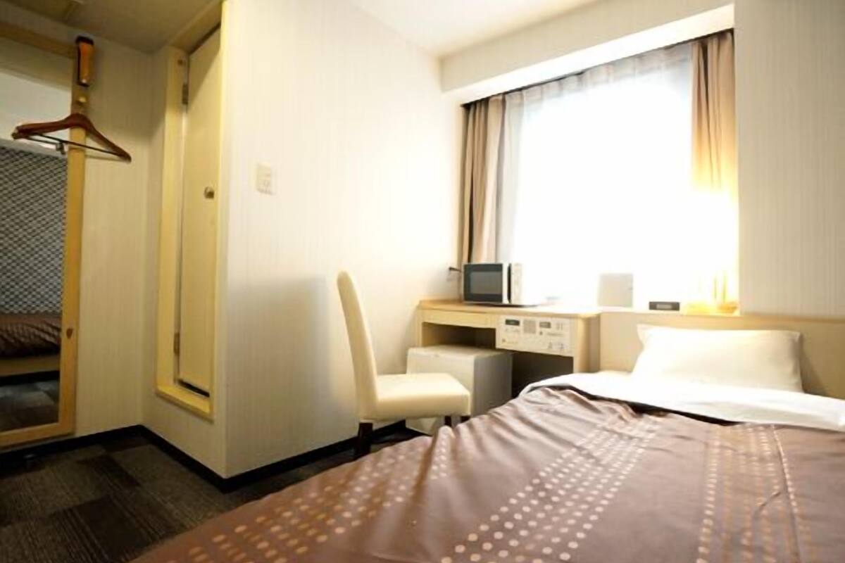 A hotel room with a bed, a desk with a computer, a chair, a TV, and a window with curtains.